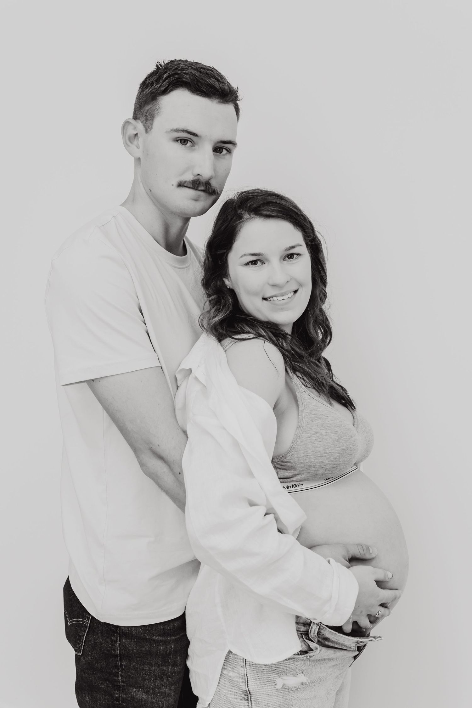 A romantic black and white maternity photo of a couple embracing against a light background.