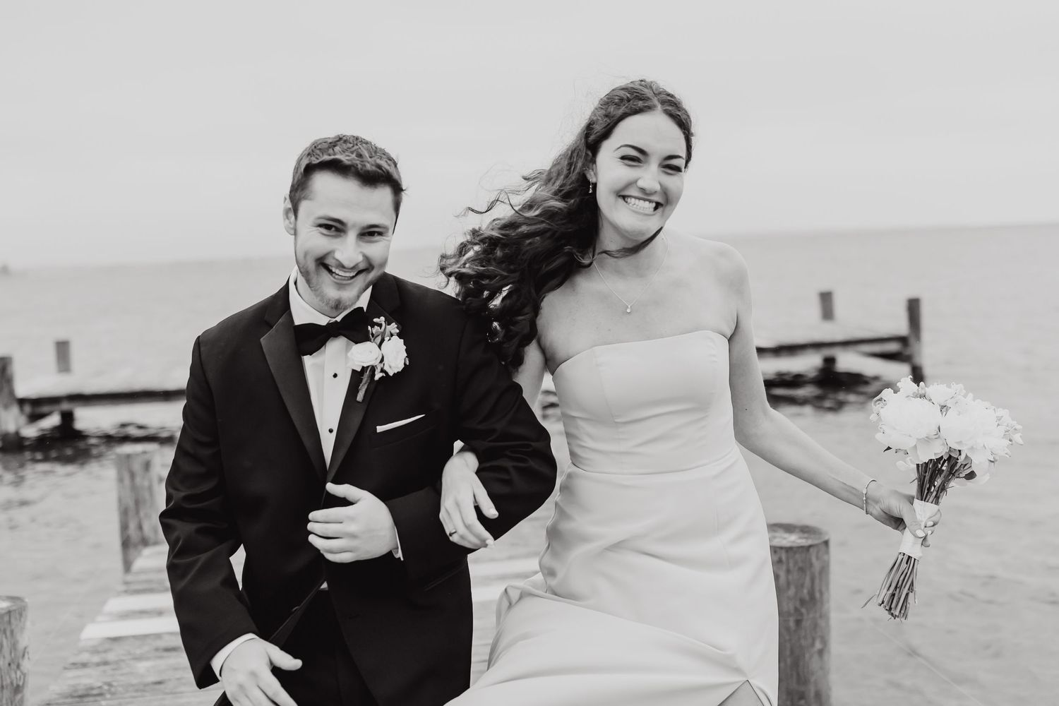 A joyful bride and groom share a candid moment together on a dock in black and white photography.