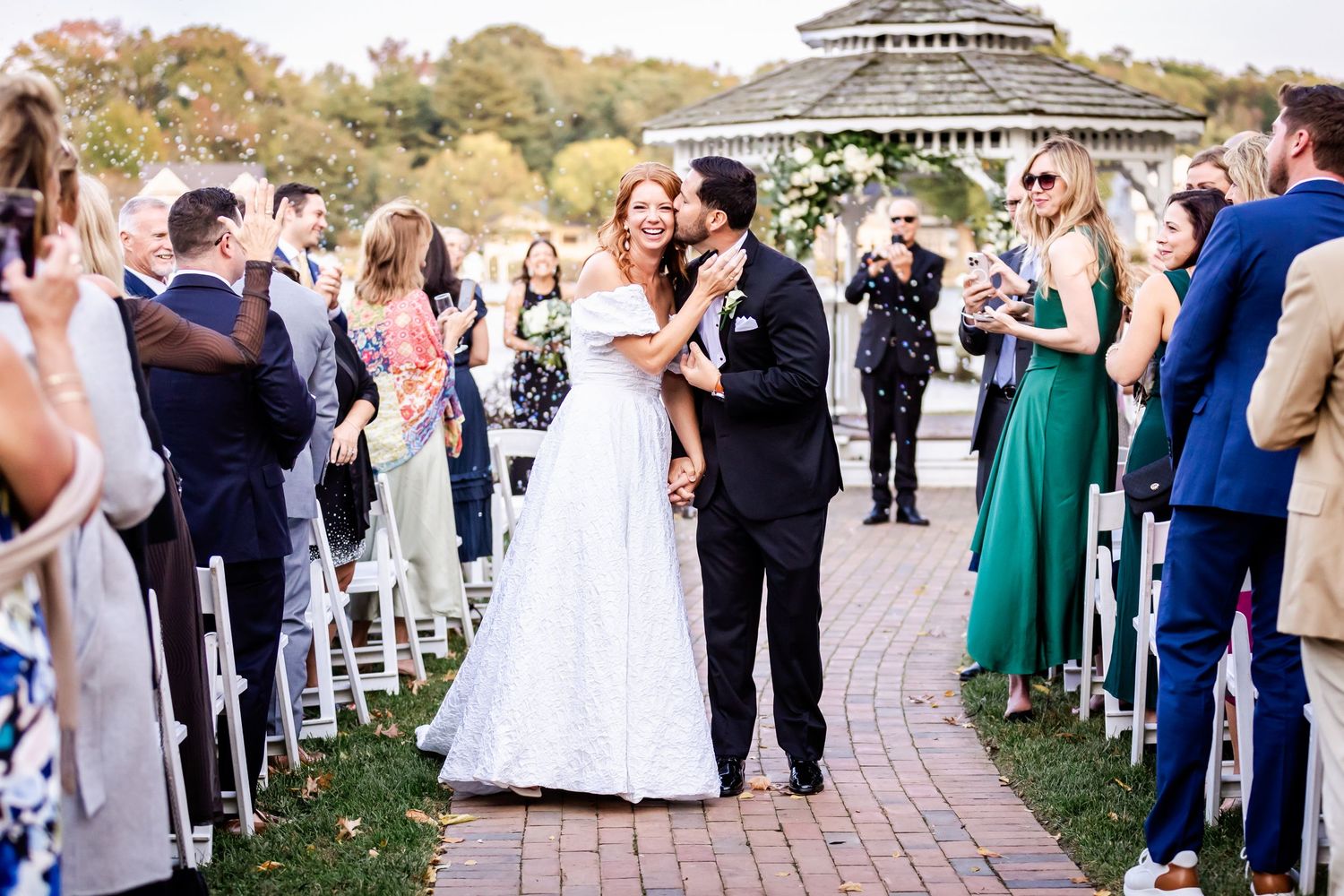 Newlywed couple shares their first kiss at outdoor wedding ceremony surrounded by guests on paved walkway near gazebo.