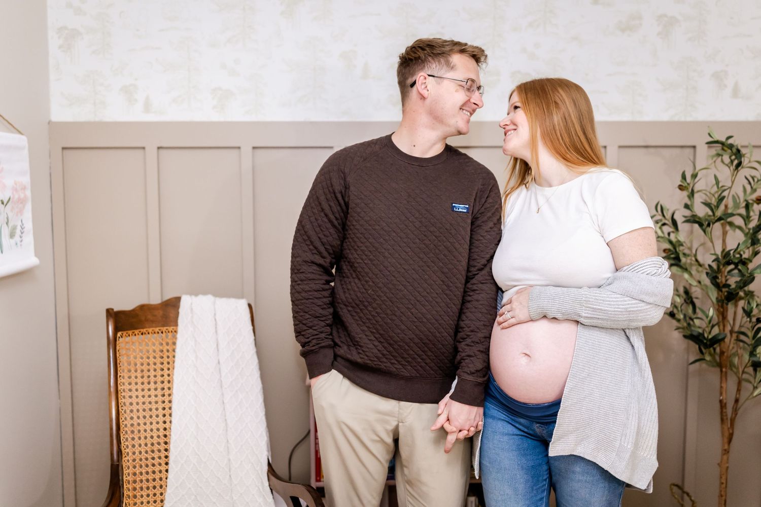 Expecting parents share tender moment together in minimalist nursery with rattan furniture.