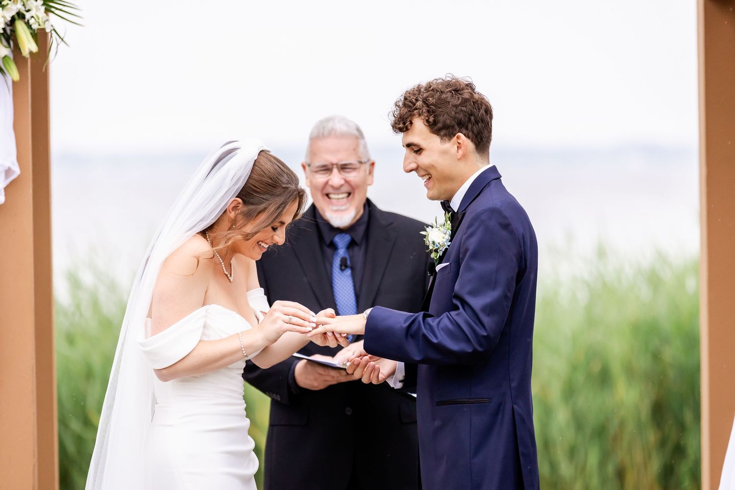 A wedding ceremony takes place under a wooden pergola with guests watching.