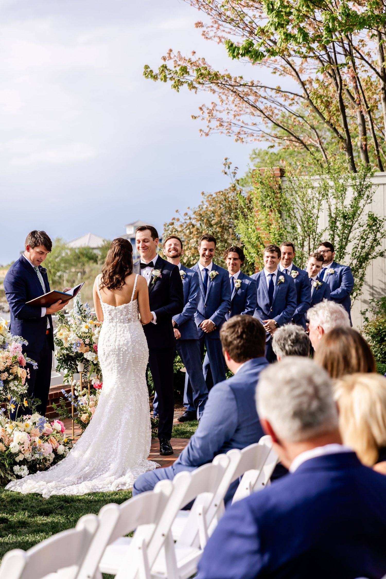 Outdoor wedding ceremony under blooming trees with guests seated on white chairs.