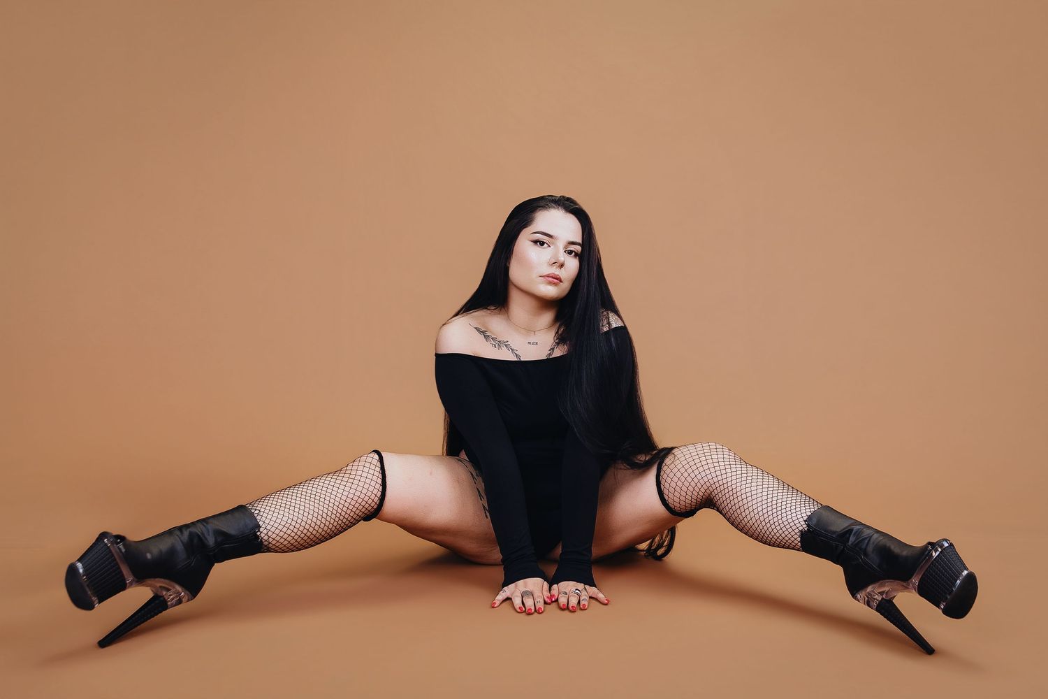 Young woman in black outfit and fishnet stockings poses in splits on beige studio background.