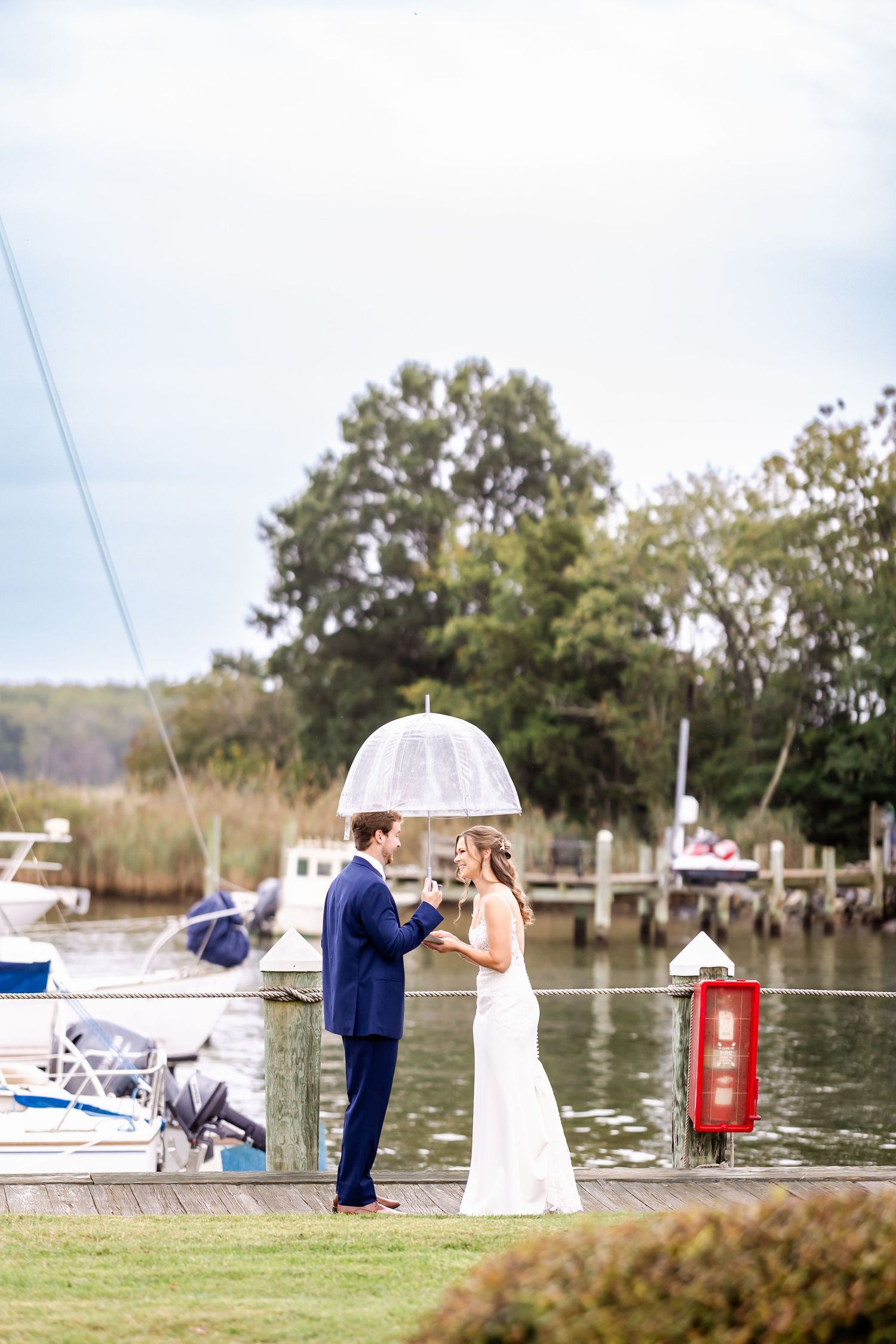 Couple in formal attire poses for romantic photos on a dock by the water with boats in the background.