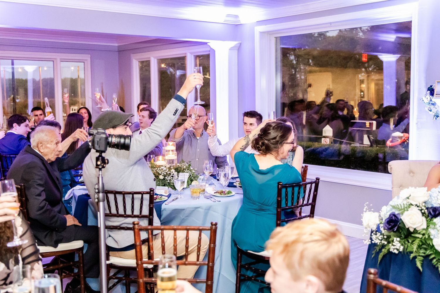 Wedding reception guests raise their glasses in celebration at an elegantly decorated venue with purple lighting.