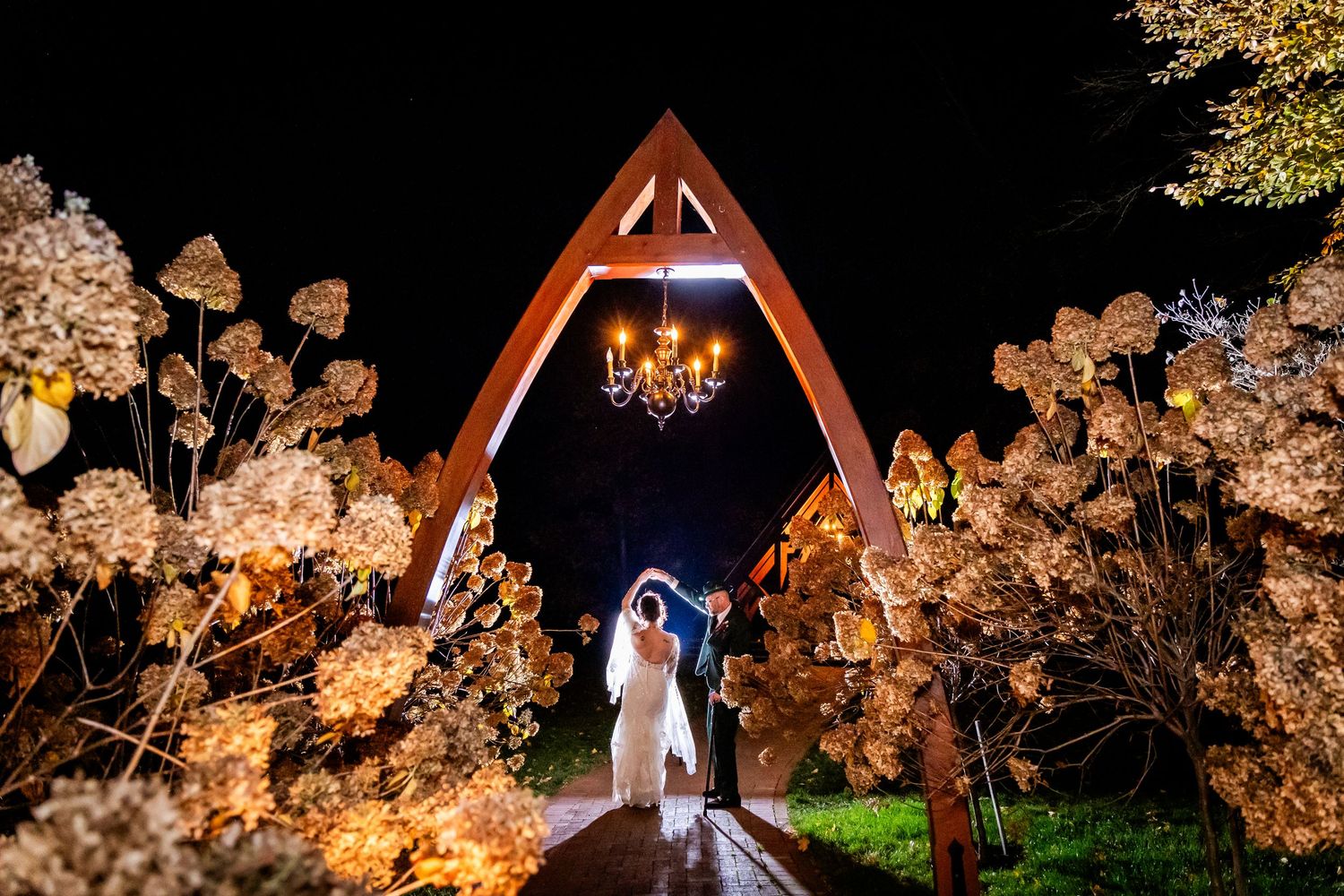 A nighttime wedding photo under an A-frame chapel with glowing chandelier and dried floral decorations.