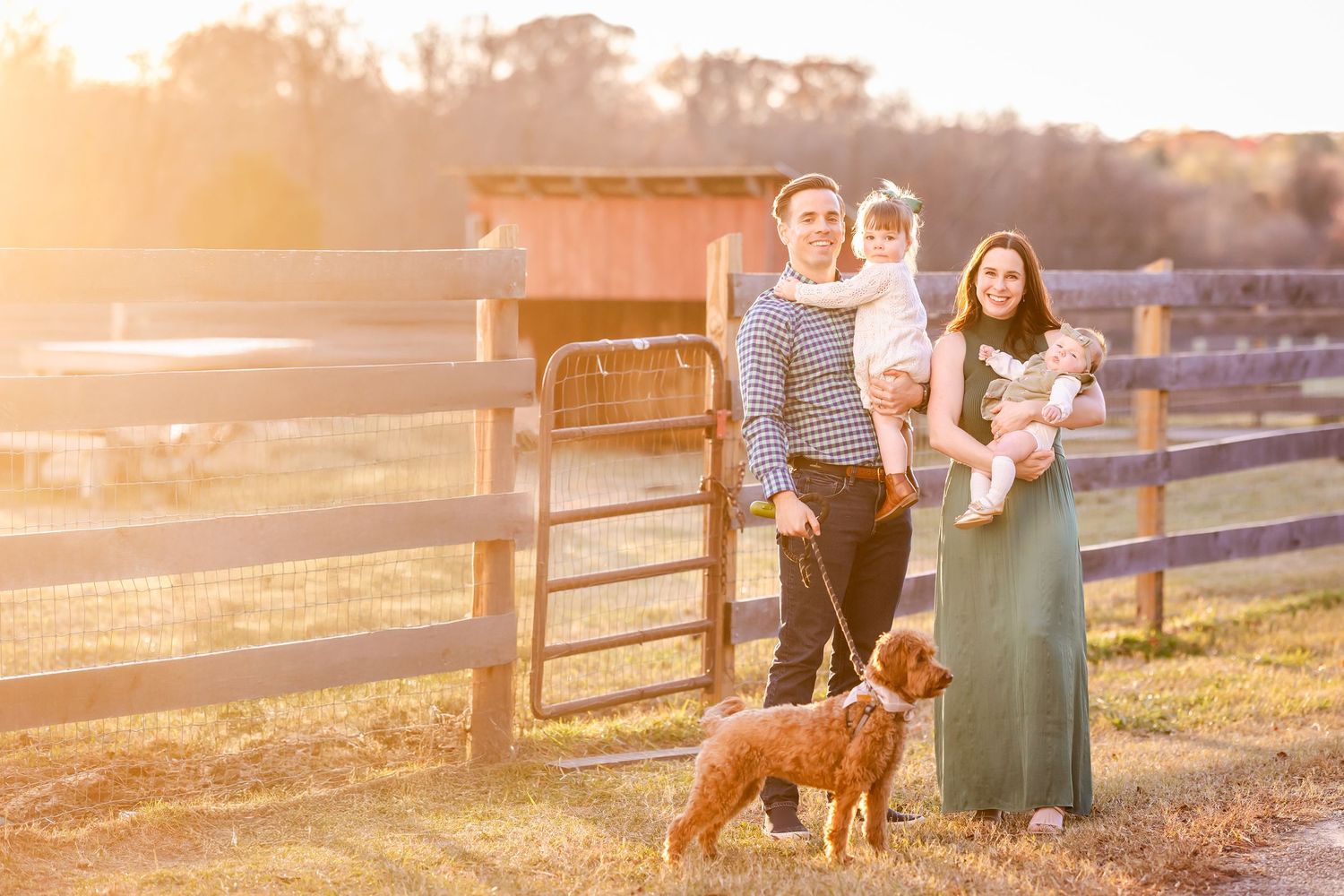 A family and their brown dog stand by a wooden fence at sunset on a rural farm property.