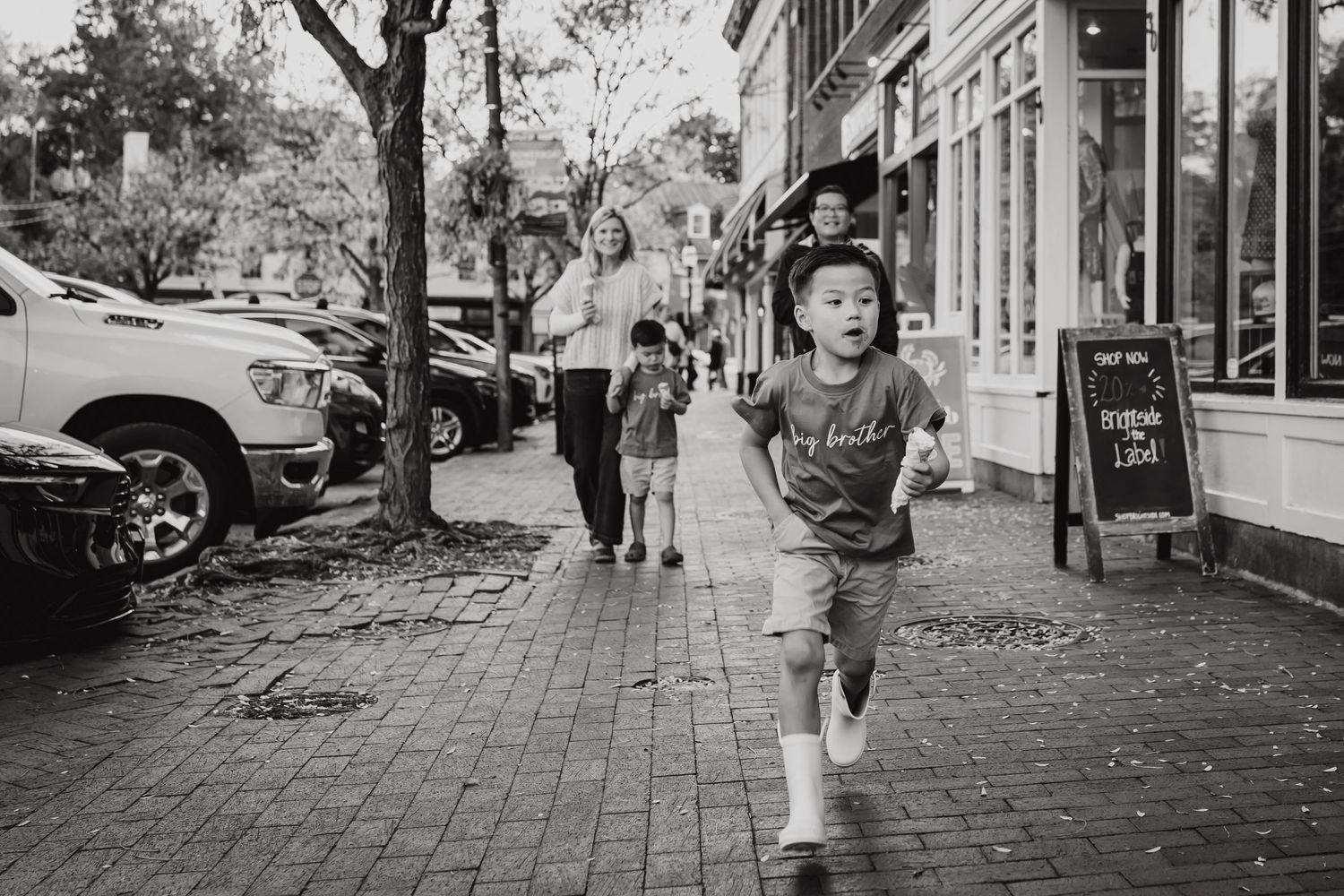 A child runs joyfully down a brick sidewalk past storefronts and parked cars in a black and white photograph.