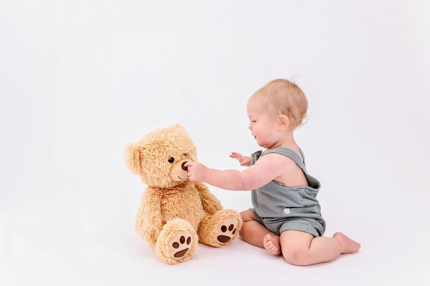 Baby in gray onesie plays with large teddy bear on white background in three sequential photos.