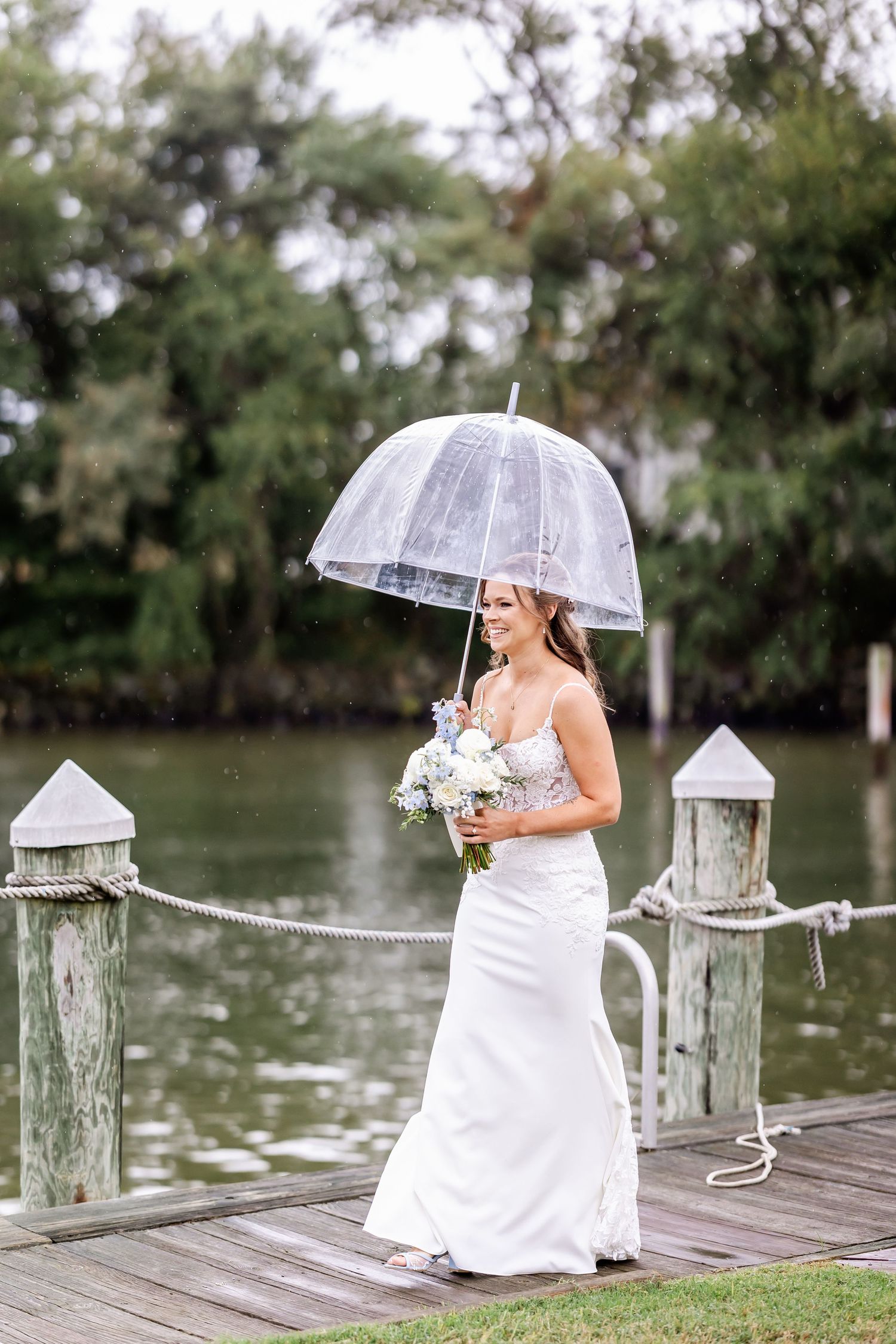A bride in a white dress holds a bouquet while standing on a dock with a clear umbrella in the rain.