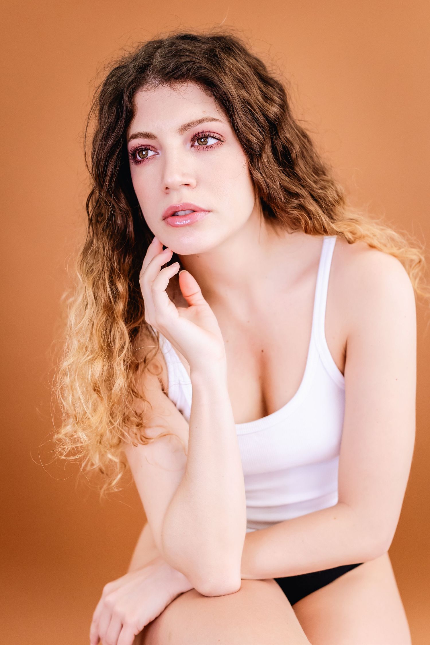 Studio portrait of a person with long wavy ombre hair wearing a white tank top against an orange background.