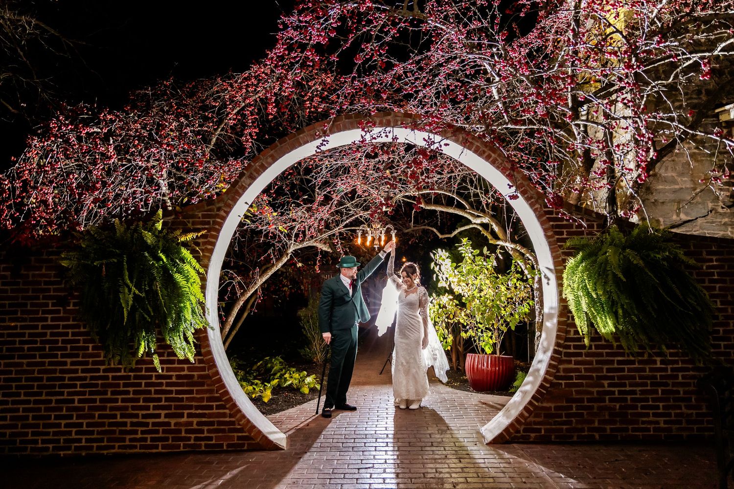 A romantic nighttime wedding photo under a circular archway with illuminated cherry blossoms.