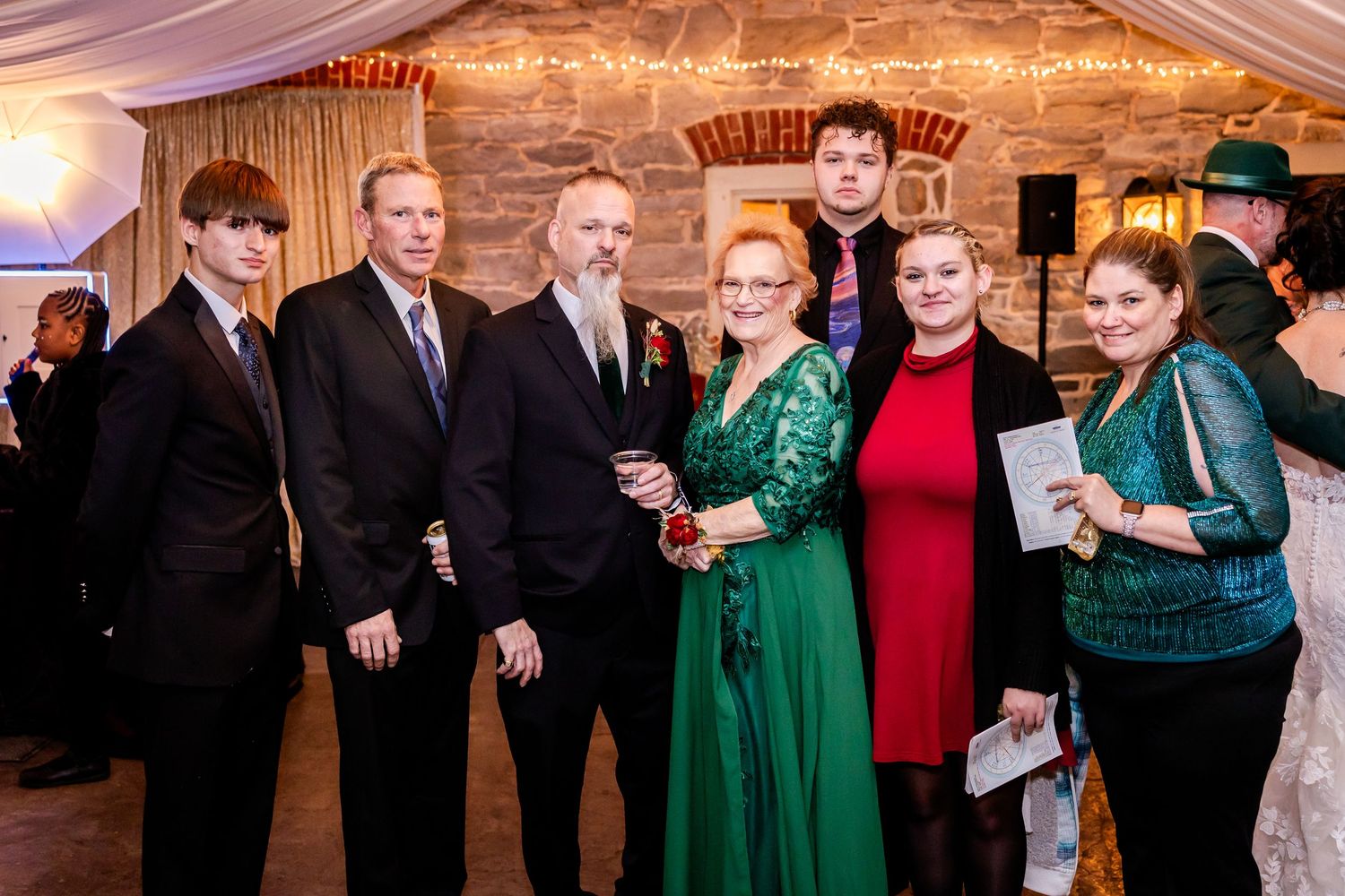 A group poses for a formal photo at an indoor wedding reception with brick wall backdrop.