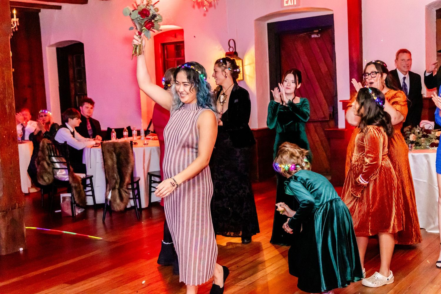 Wedding guests celebrating on a dance floor under pink lighting with exposed wooden beams overhead.