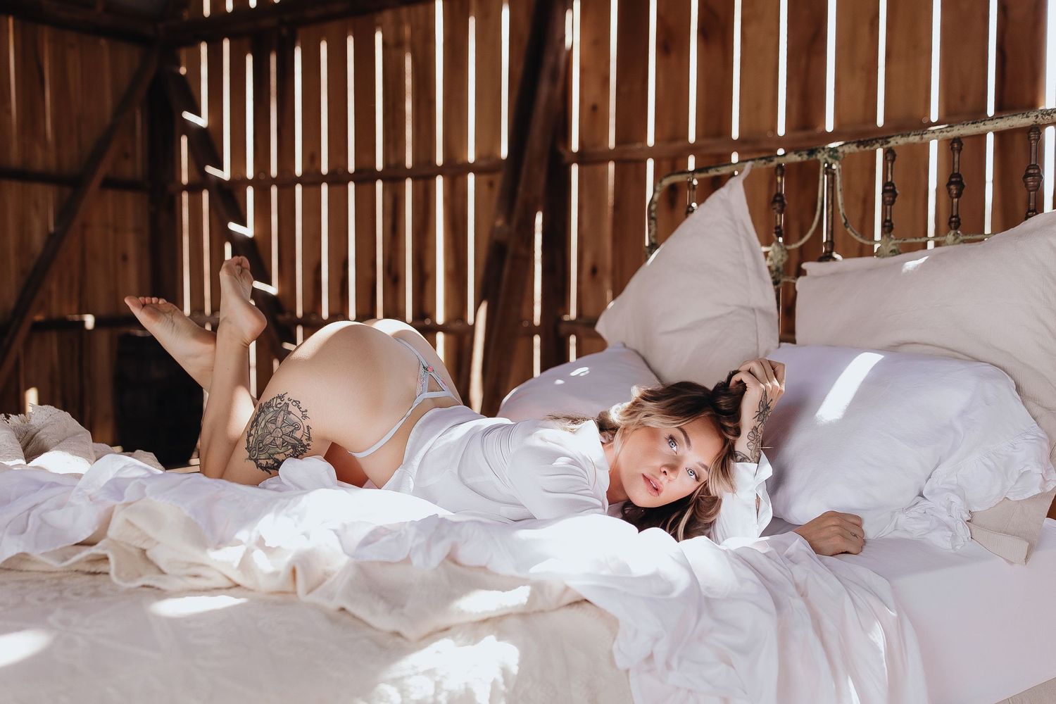 Woman in white shirt lying on a bed with white bedding in a rustic barn setting with wooden walls.