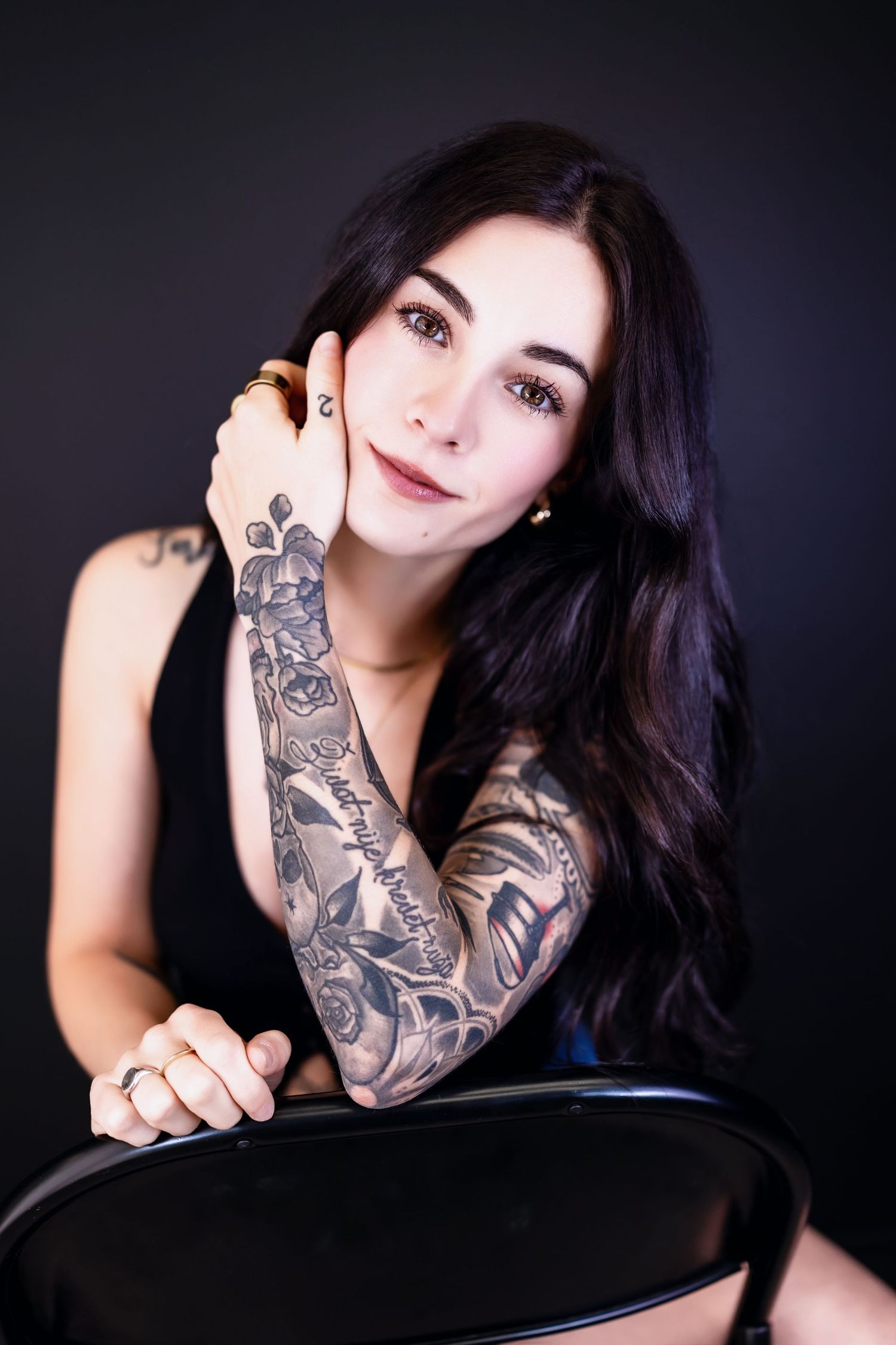 Young woman with long dark hair and full sleeve tattoo poses confidently against a dark background.