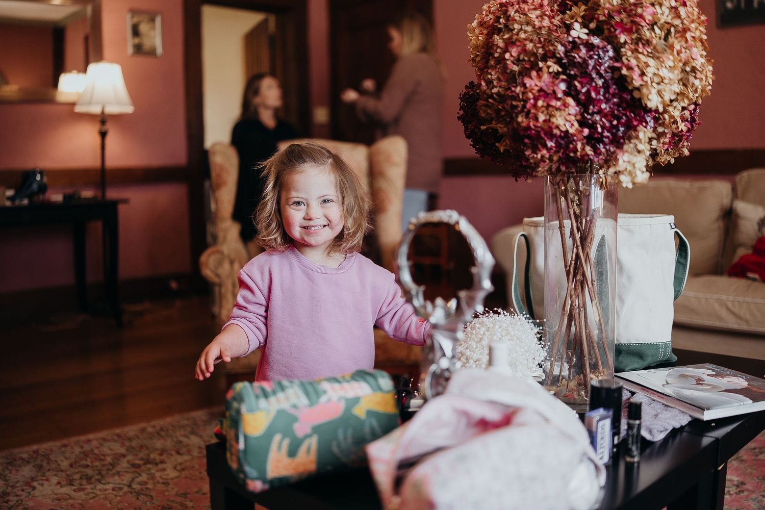 Young toddler girl in pink sweater smiles beside wrapped gifts and dried flower arrangement in cozy living room.