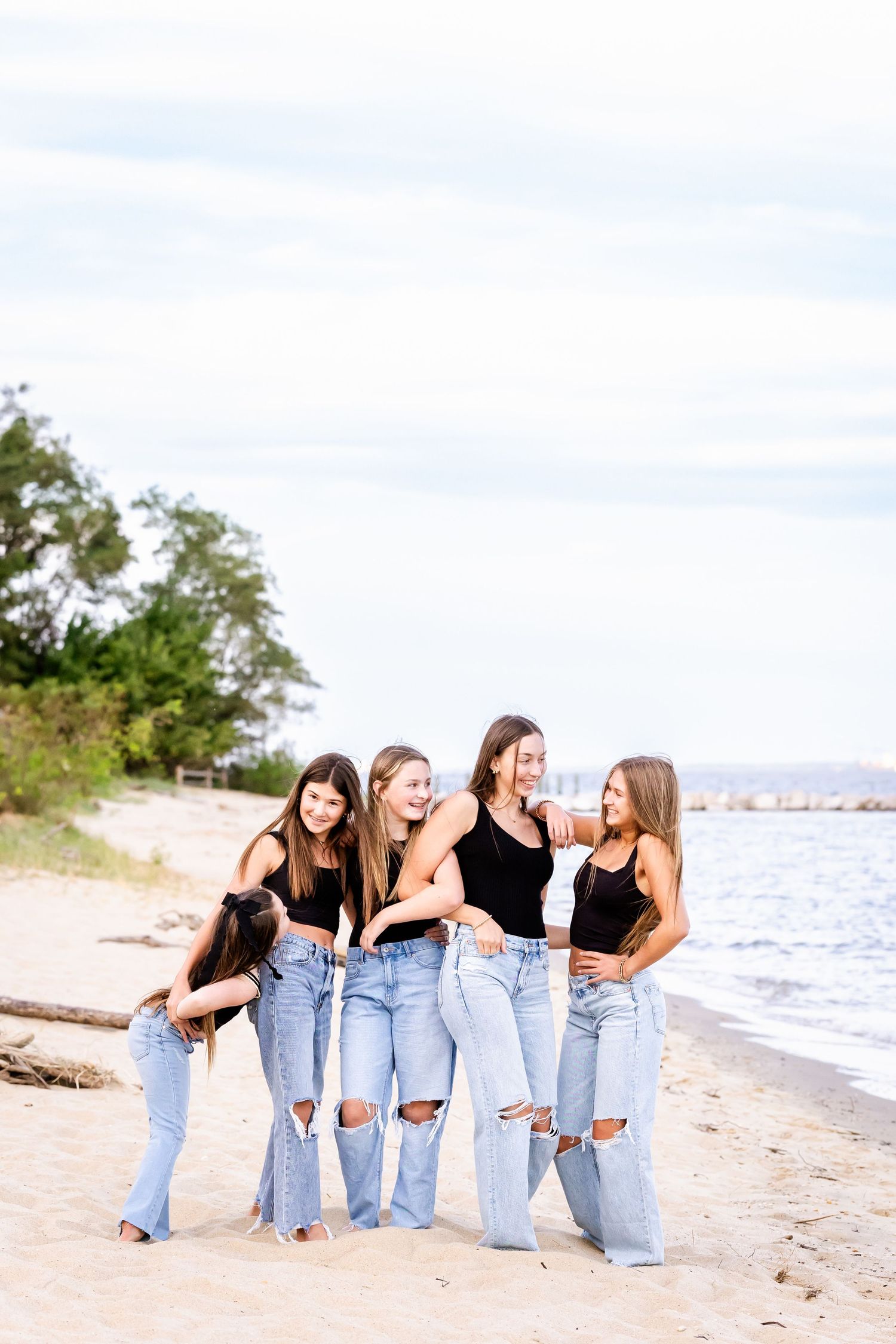 Group of friends wearing matching ripped jeans pose together on a sunny beach with ocean waves in the background.