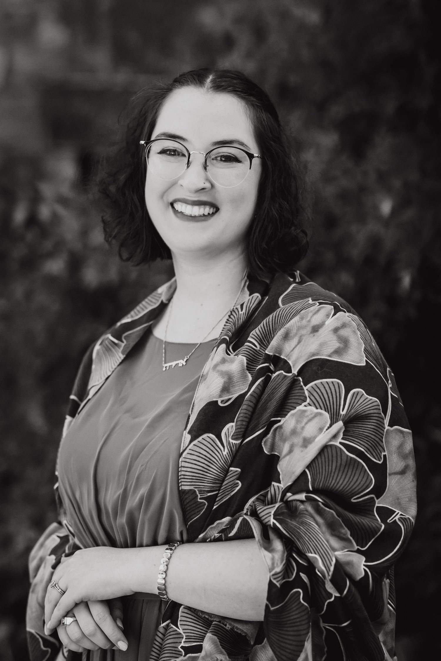 A black and white portrait of a smiling woman wearing glasses and a floral wrap dress.