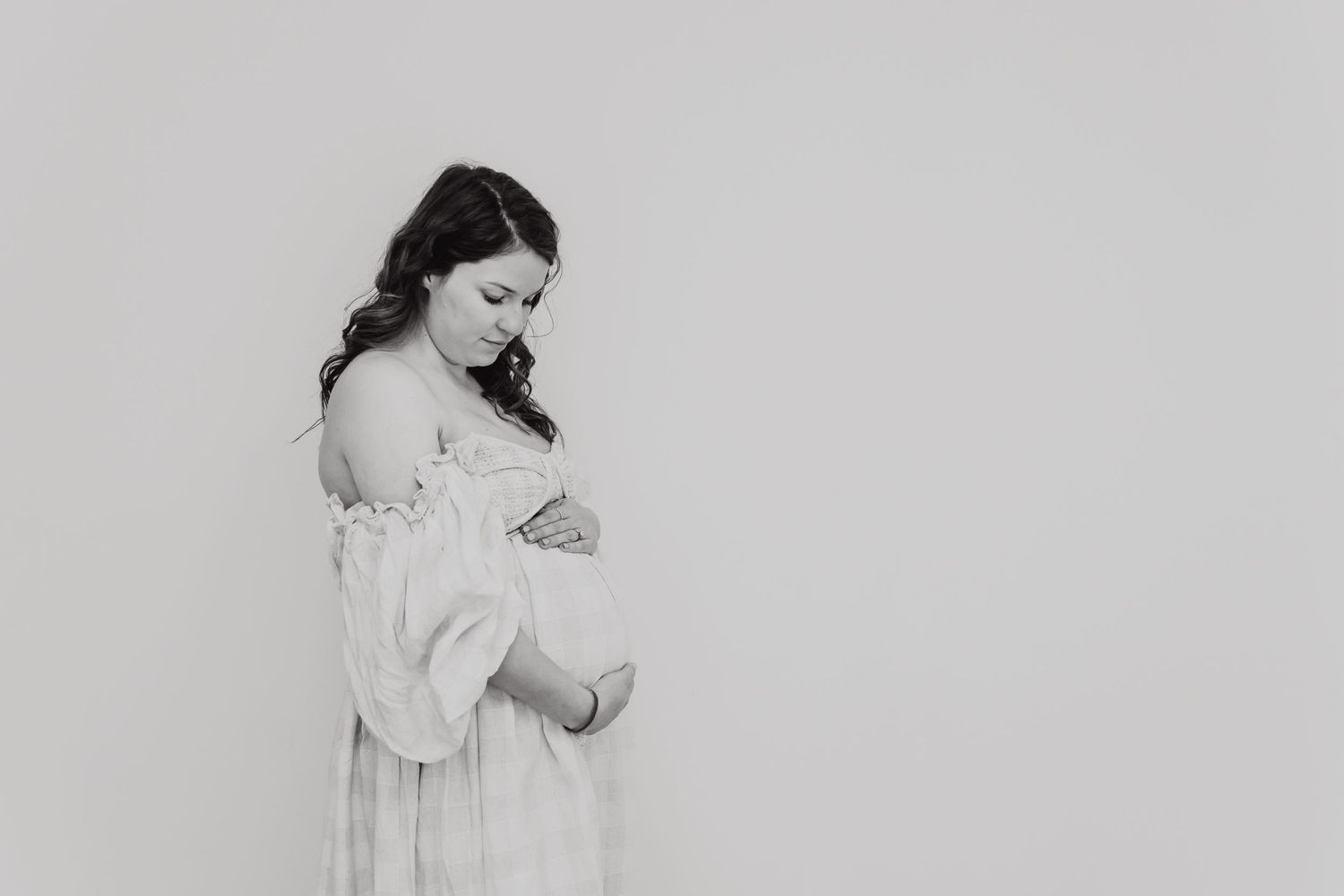 Black and white image of a woman in a white dress looking down at her pregnant belly against a plain background.