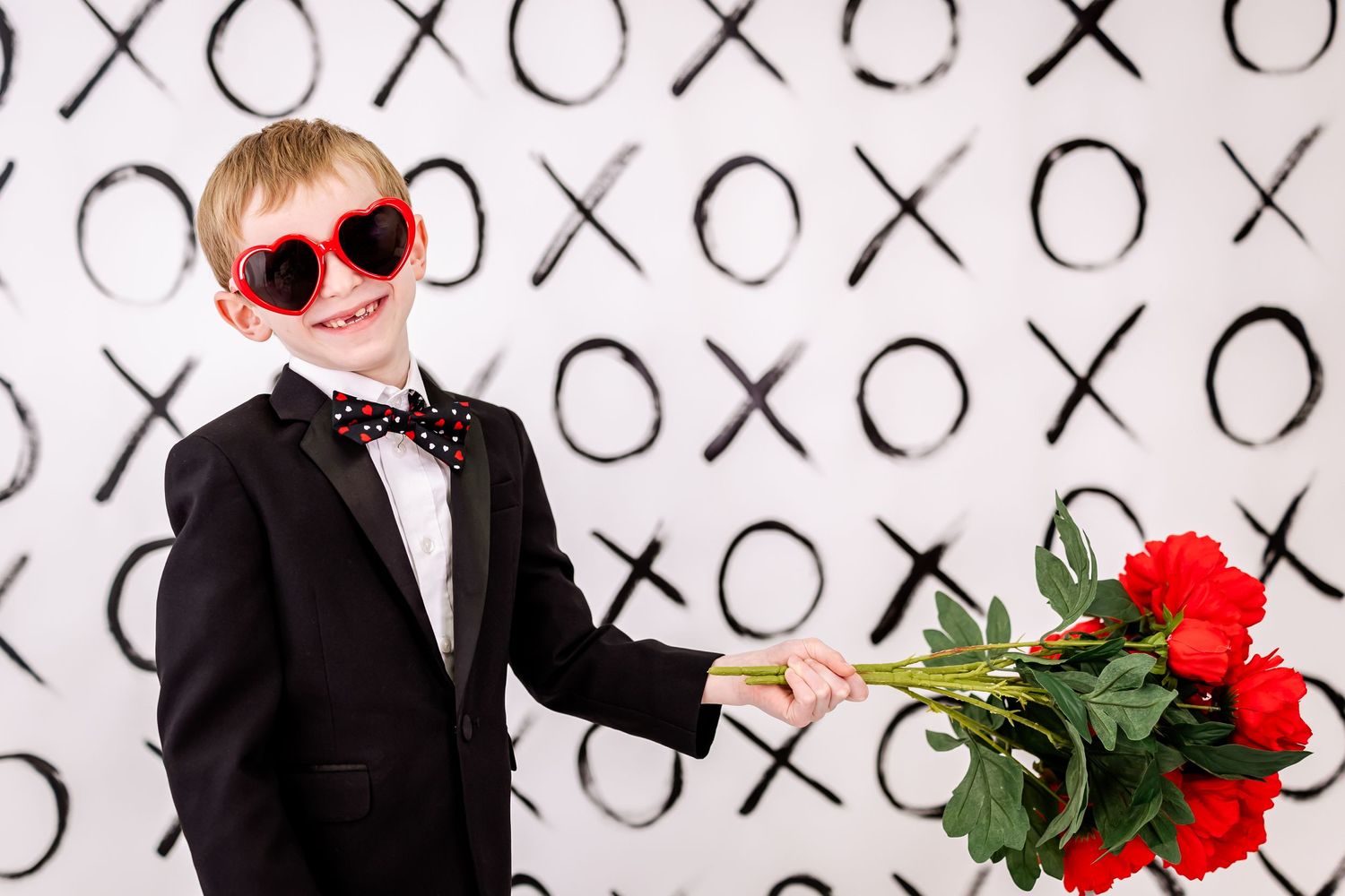 Person in tuxedo and red sunglasses holds red roses against black and white X's and O's backdrop.