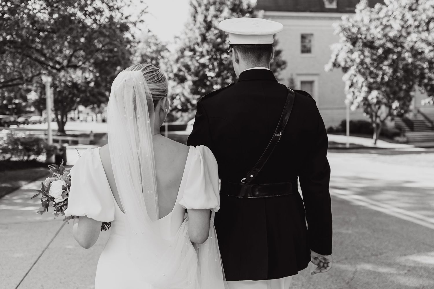 Black and white photograph capturing the rear view of a bride in a white dress and groom in military uniform walking together.