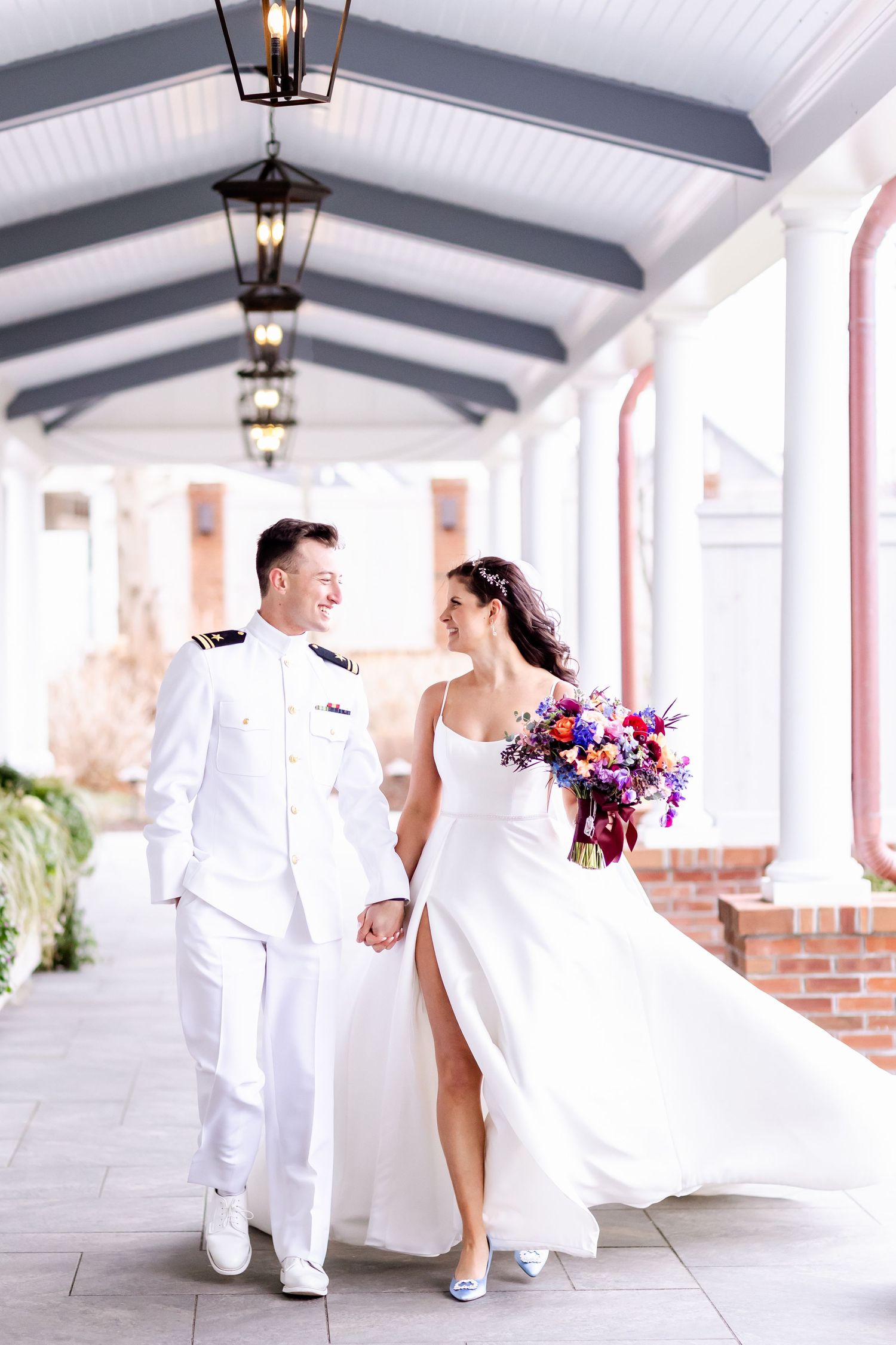 A couple in white attire shares romantic moments while walking along an elegant covered porch with lanterns overhead.