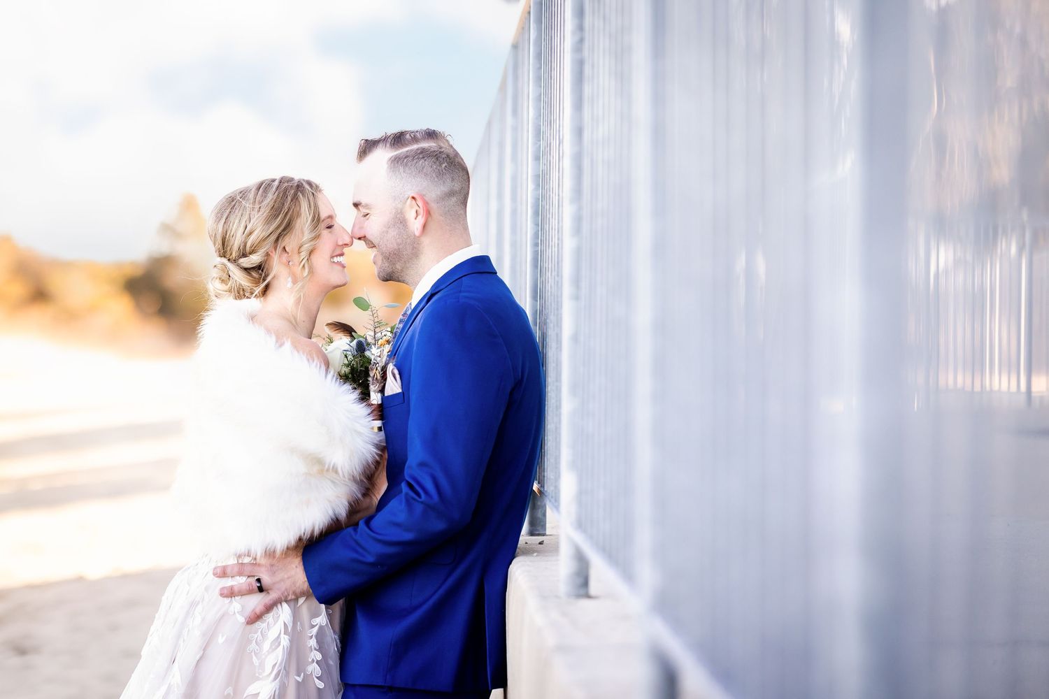 Romantic wedding couple embracing against a white wall with the bride wearing a fur wrap and groom in blue suit.