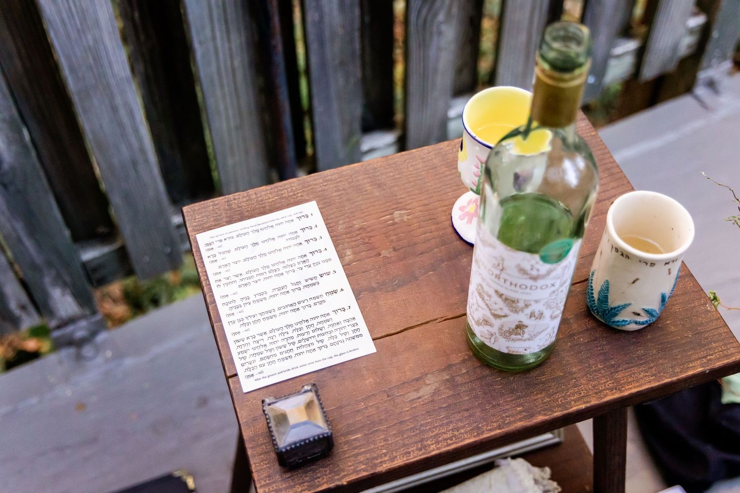 Wedding ceremony details on wooden table showing document, wine bottle and cups outdoors.