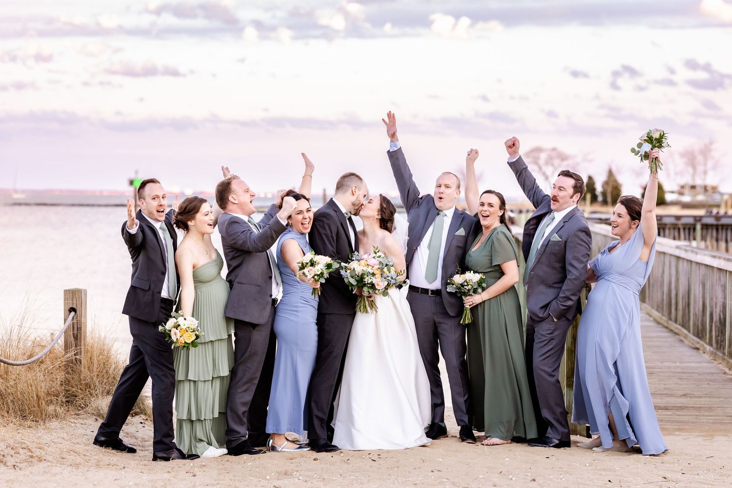 Wedding party celebrating joyfully on wooden boardwalk by water with raised arms at sunset.