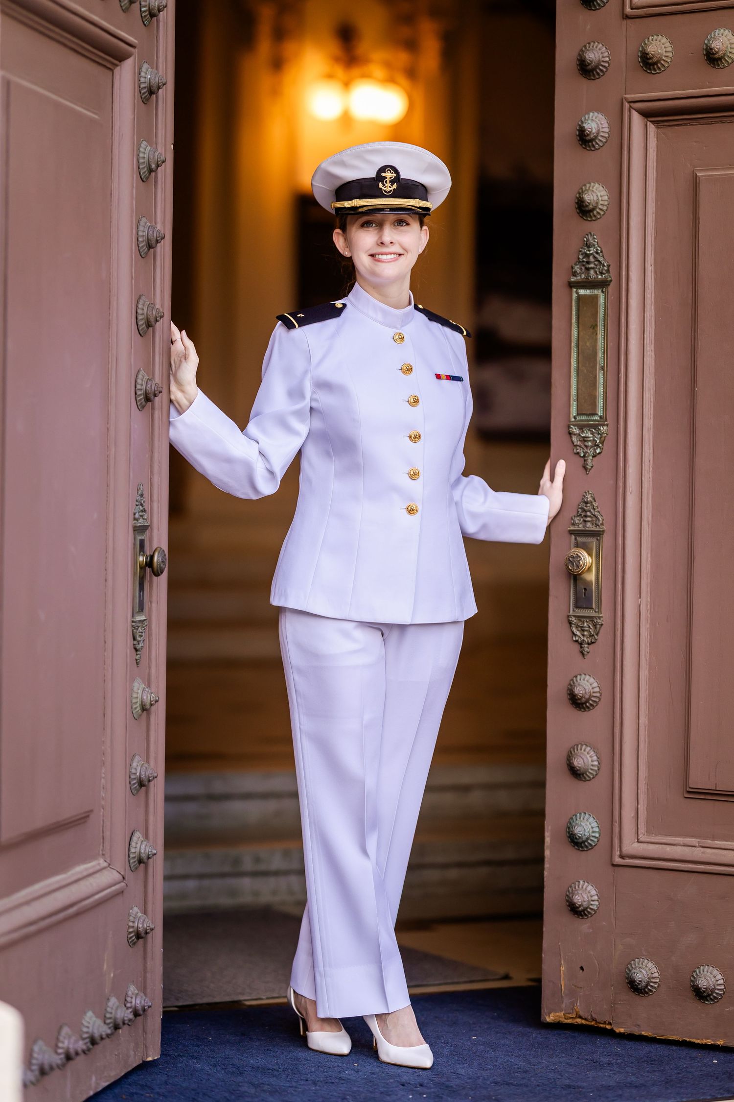 Officer in formal white military dress uniform stands elegantly between ornate wooden doors.