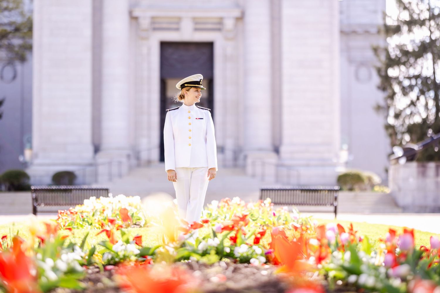 A service member in white uniform stands near colorful tulip gardens in front of a white building.