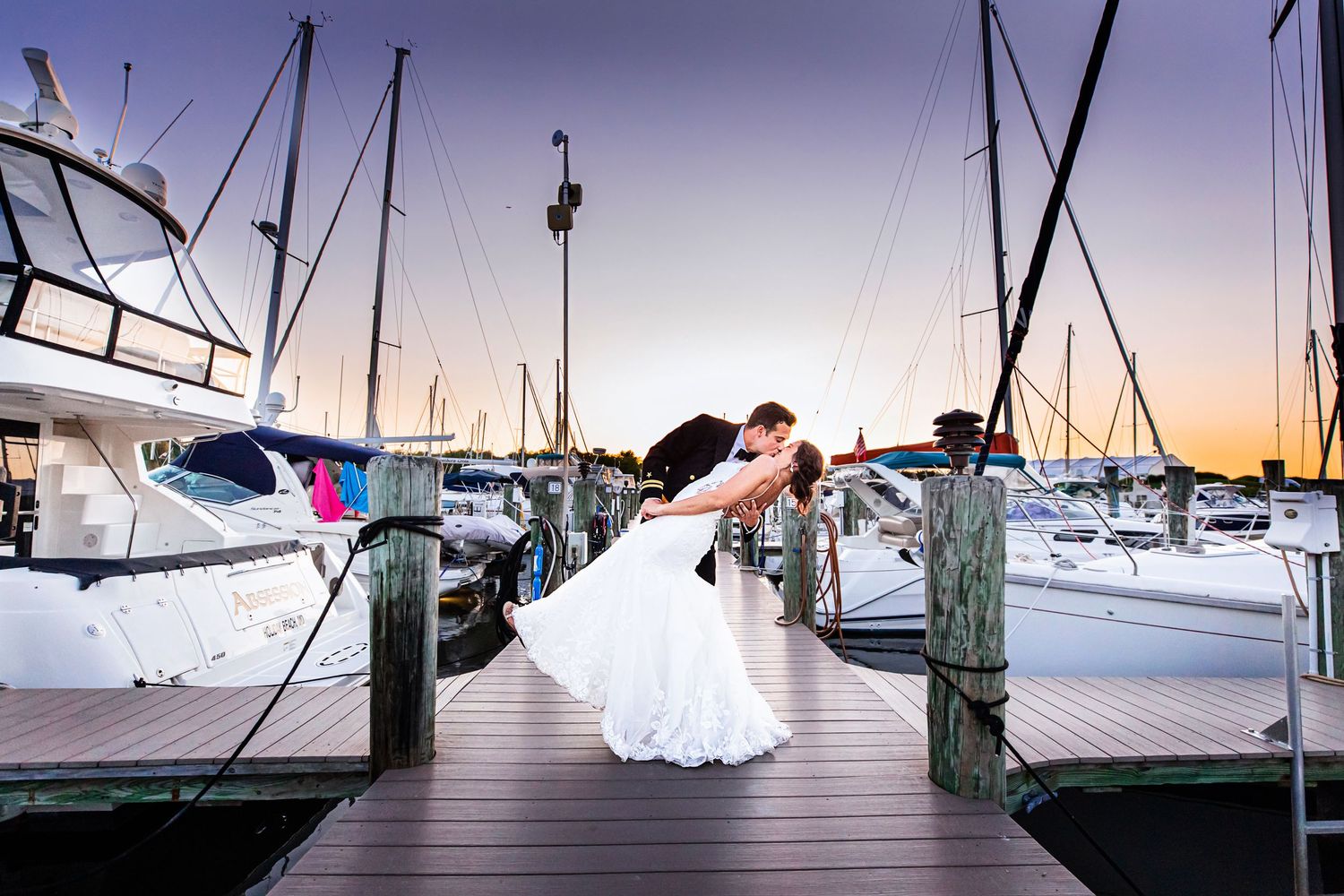 Sailboats docked at marina during sunset with wooden pier stretching across tranquil water.