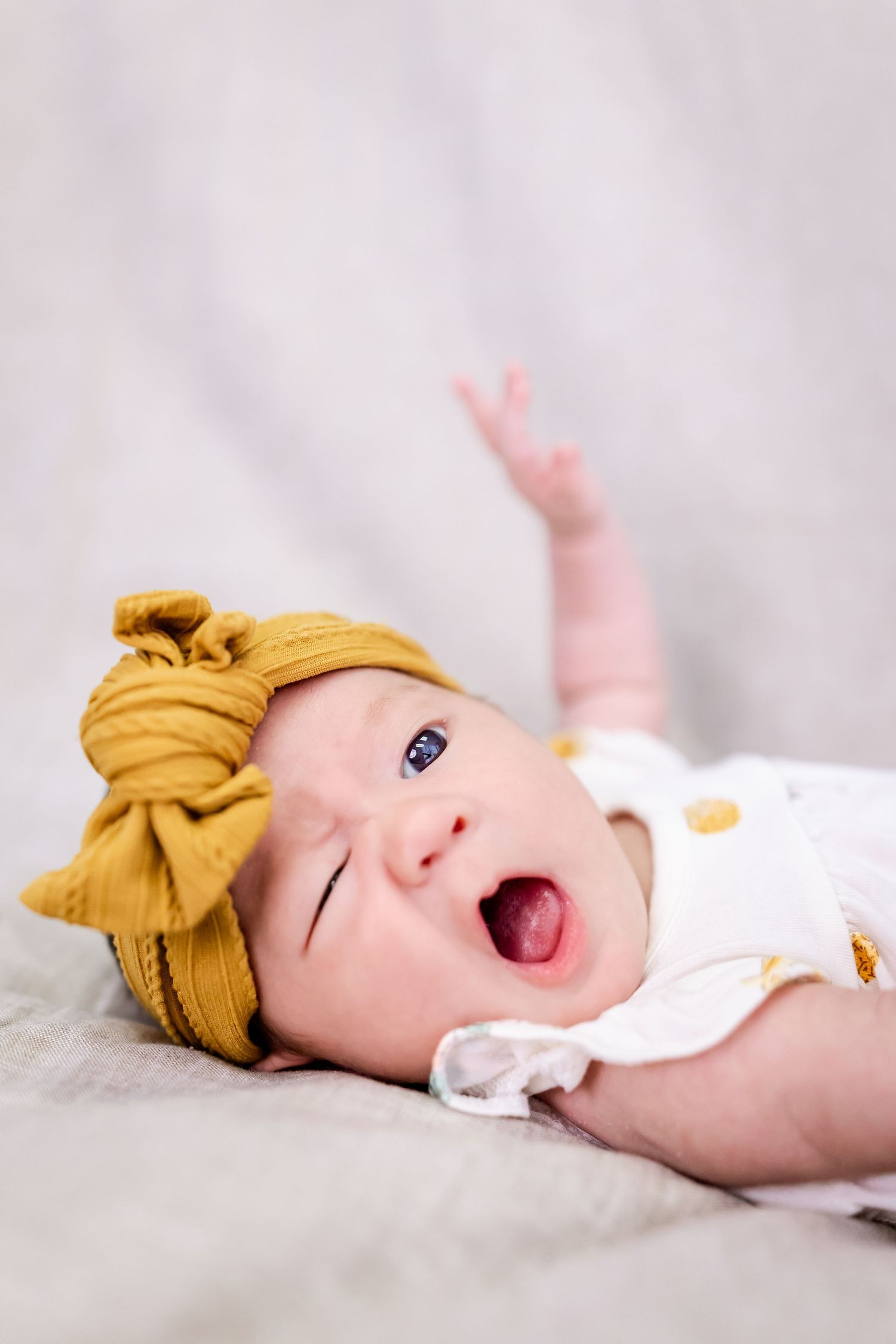Happy baby with yellow bow headband laughing and playing on light gray blanket.