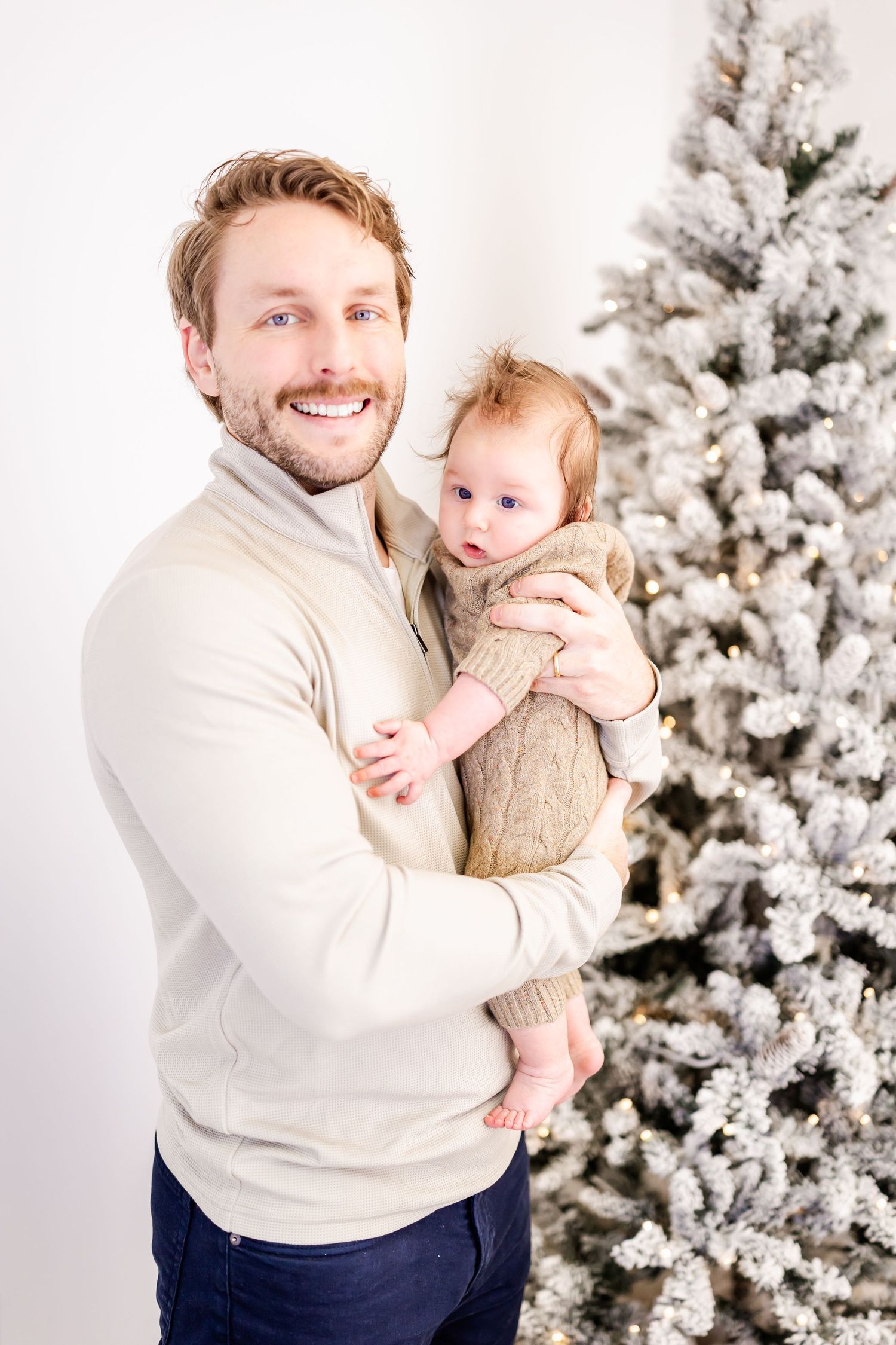 A man in a white sweater holds a baby in front of a snow-covered Christmas tree.