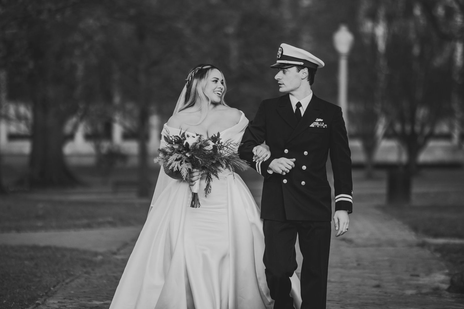 A black and white photo of a military wedding couple walking together on a tree-lined path with the bride holding a bouquet.