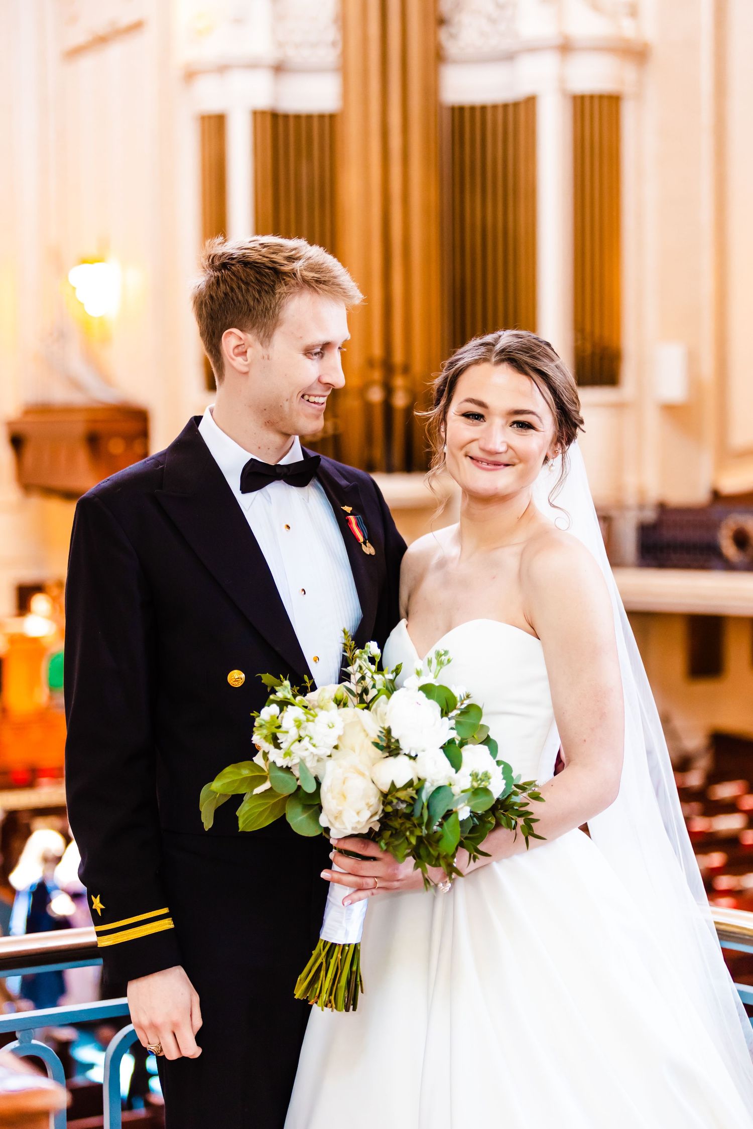 A happy couple in wedding attire smiles together while holding a white and green floral bouquet in an elegant venue.