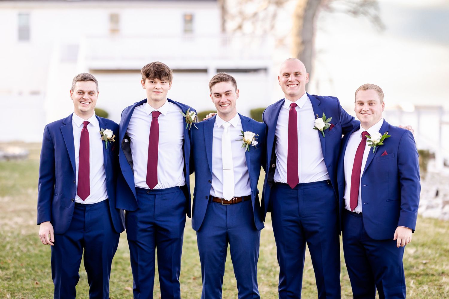 Groomsmen in navy suits and red ties pose together outdoors in a casual lineup.