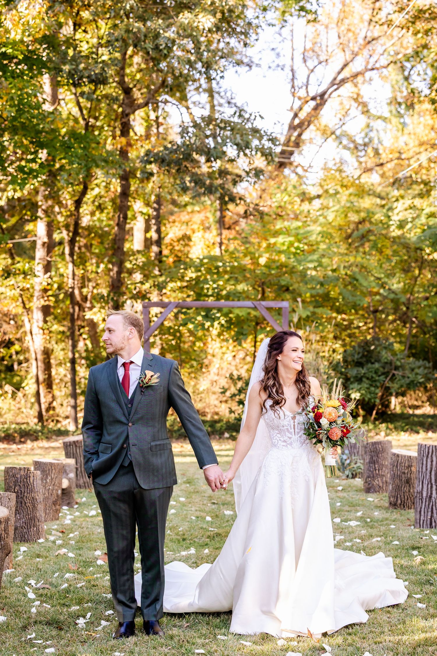 Wedding couple poses playfully on rustic wooden stumps in autumn forest setting with golden sunlight.