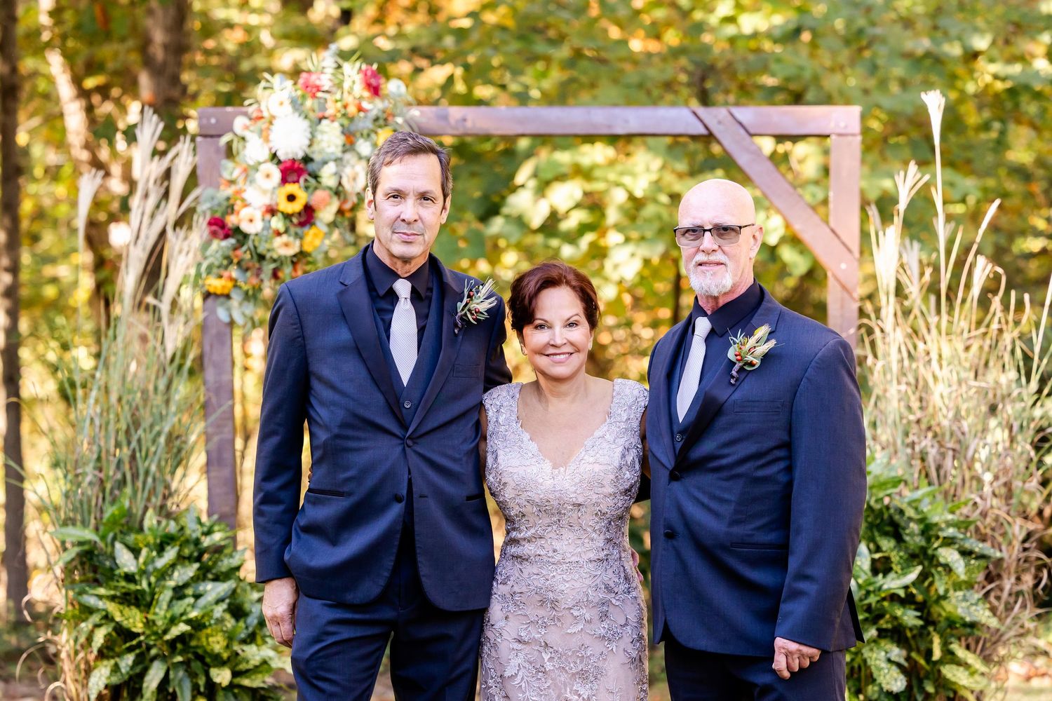 Wedding group stands together in front of wooden arch with fall trees in background.