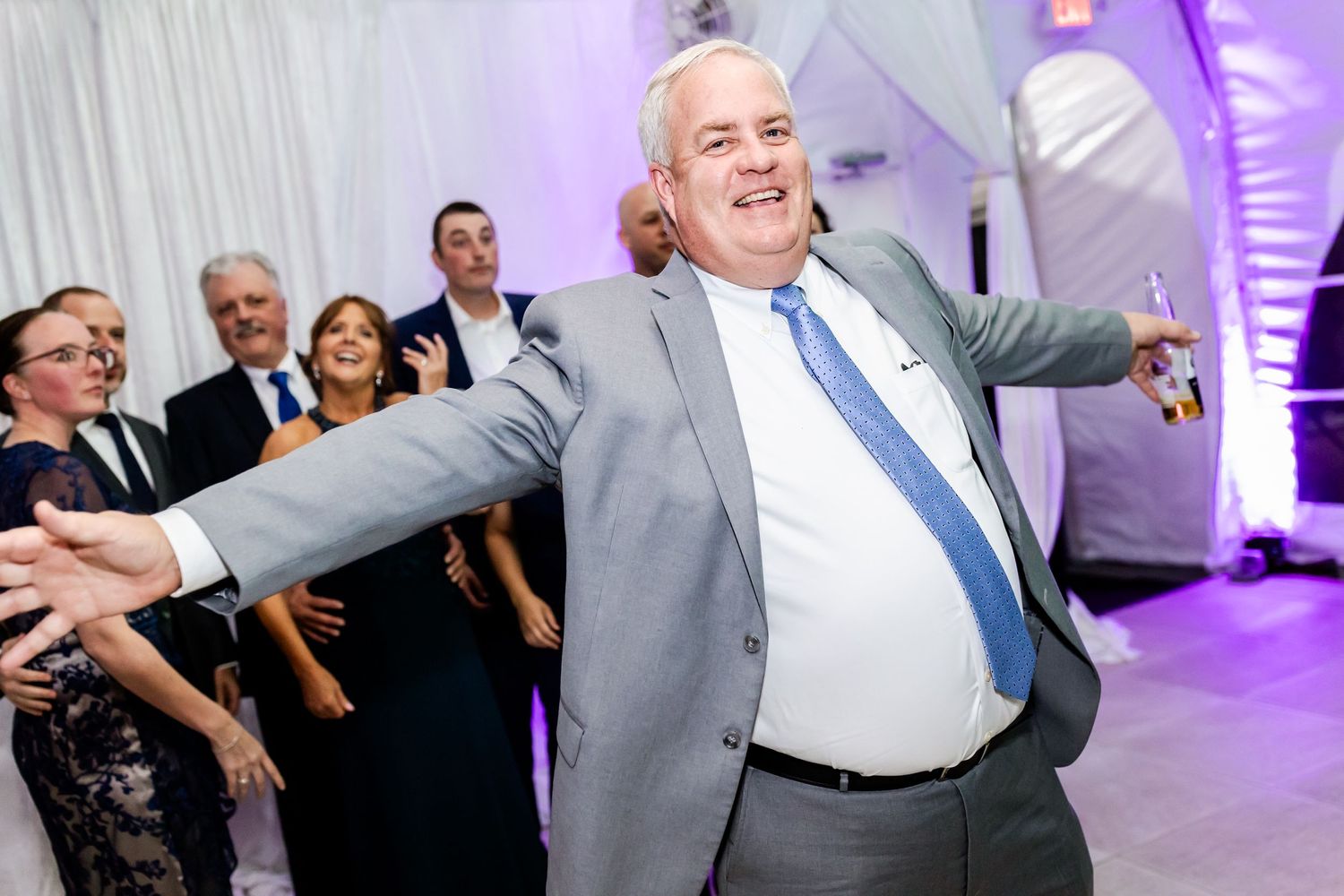 Guest in grey suit dancing enthusiastically under purple-lit tent ceiling at celebration.