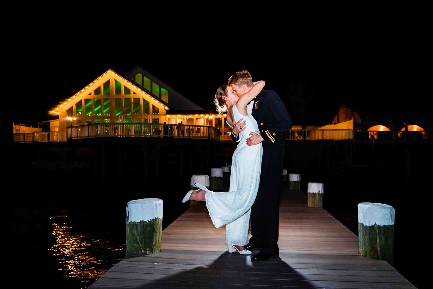 Romantic couple embraces on illuminated wooden dock at night with glowing venue in background.
