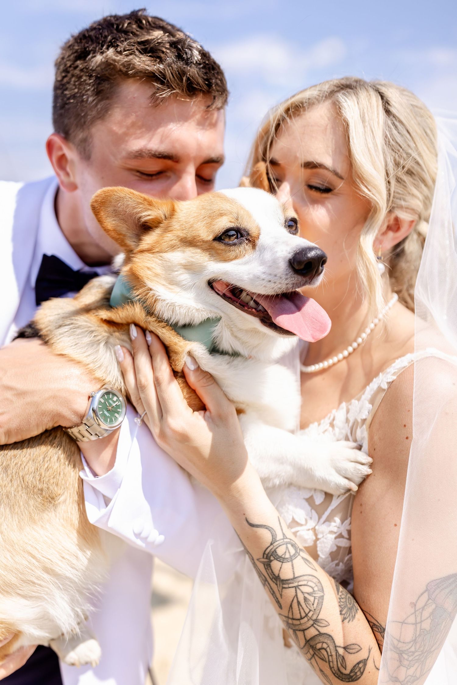 A happy Corgi dog being held and kissed by newlyweds in formal wedding attire on their special day.