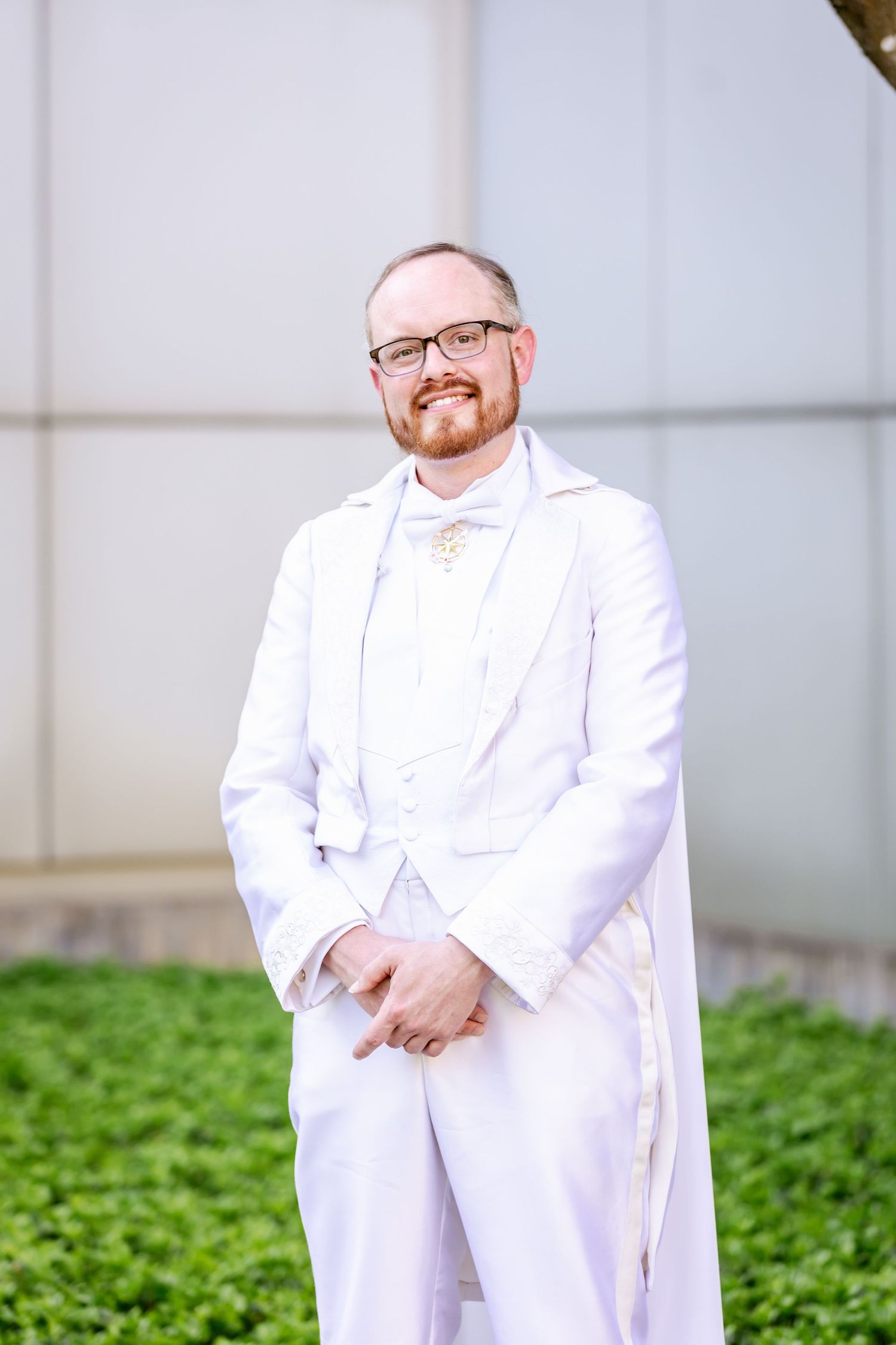 Person in crisp white wedding attire and glasses poses for a formal portrait outdoors.