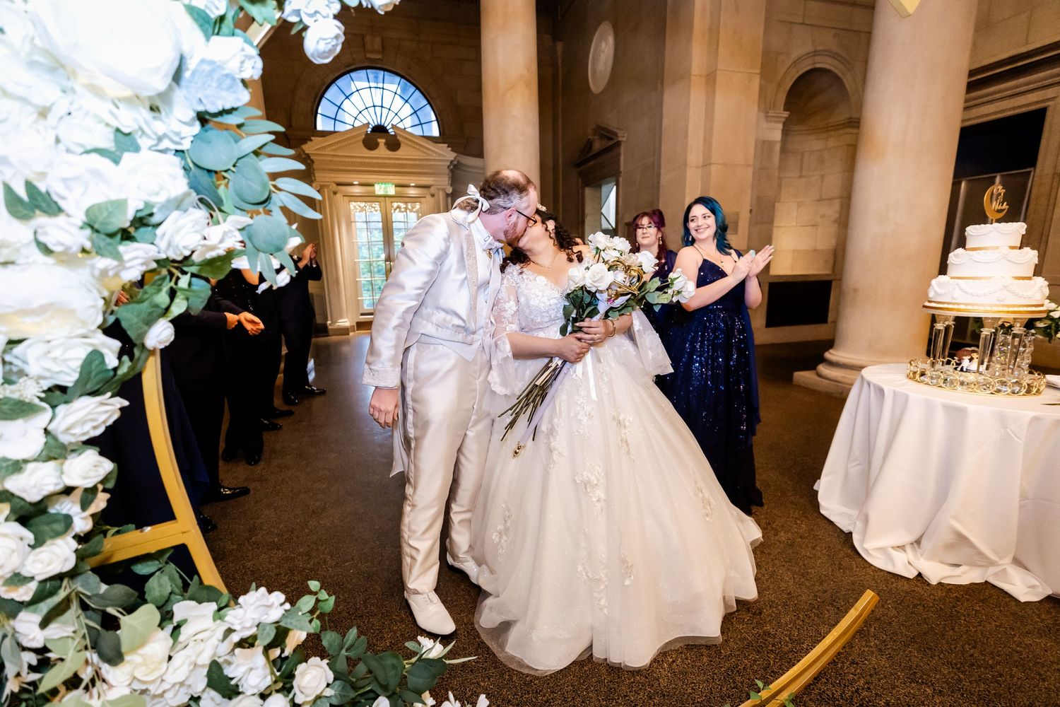 Newlyweds celebrate during reception in grand columned venue with elegant floral decorations.