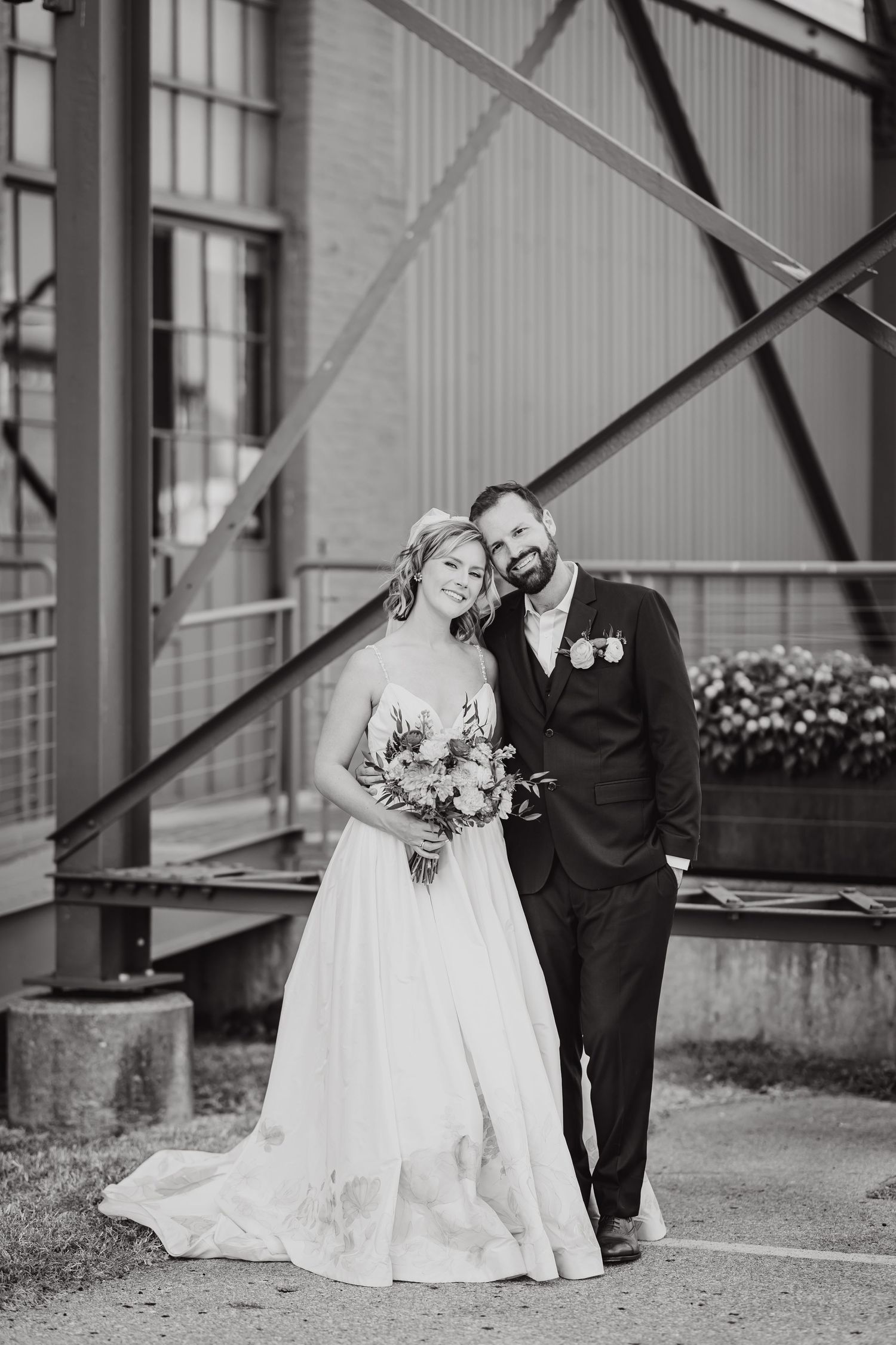 Black and white portrait of couple in wedding attire against industrial backdrop.