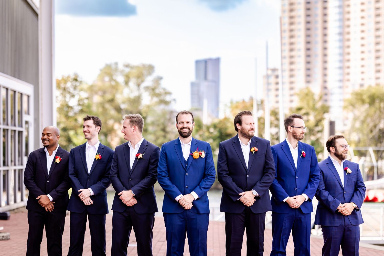 Groomsmen lined up outdoors wearing coordinated navy blue suits and smiling in front of a city backdrop.