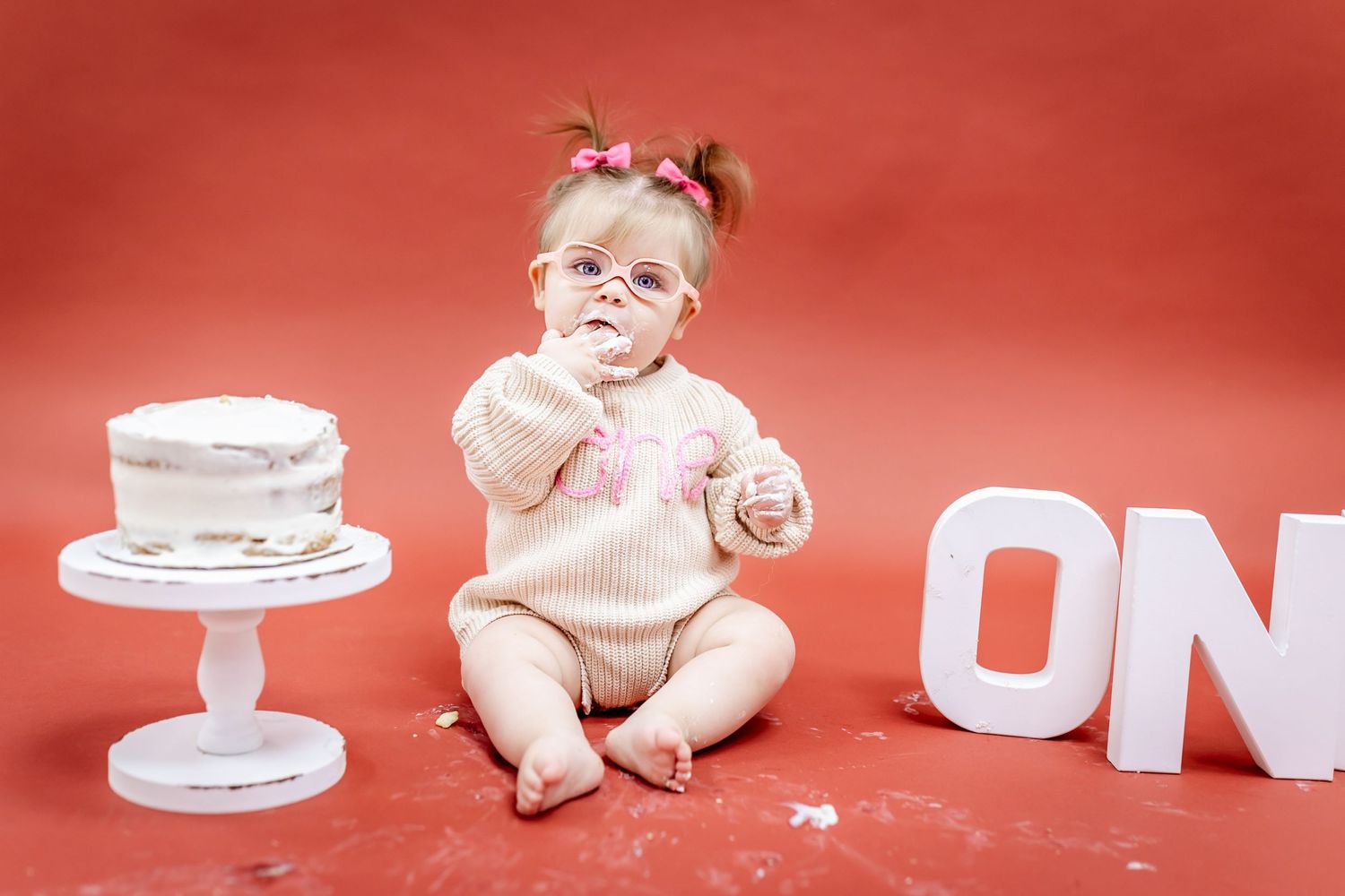 A baby in a knit sweater sits next to a white cake and decorative letters against a red background.