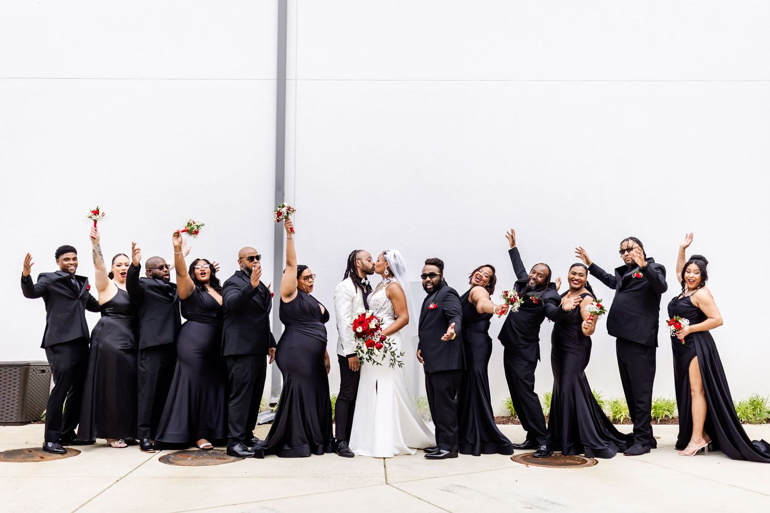 Wedding party celebrates with raised arms against white wall outdoors.