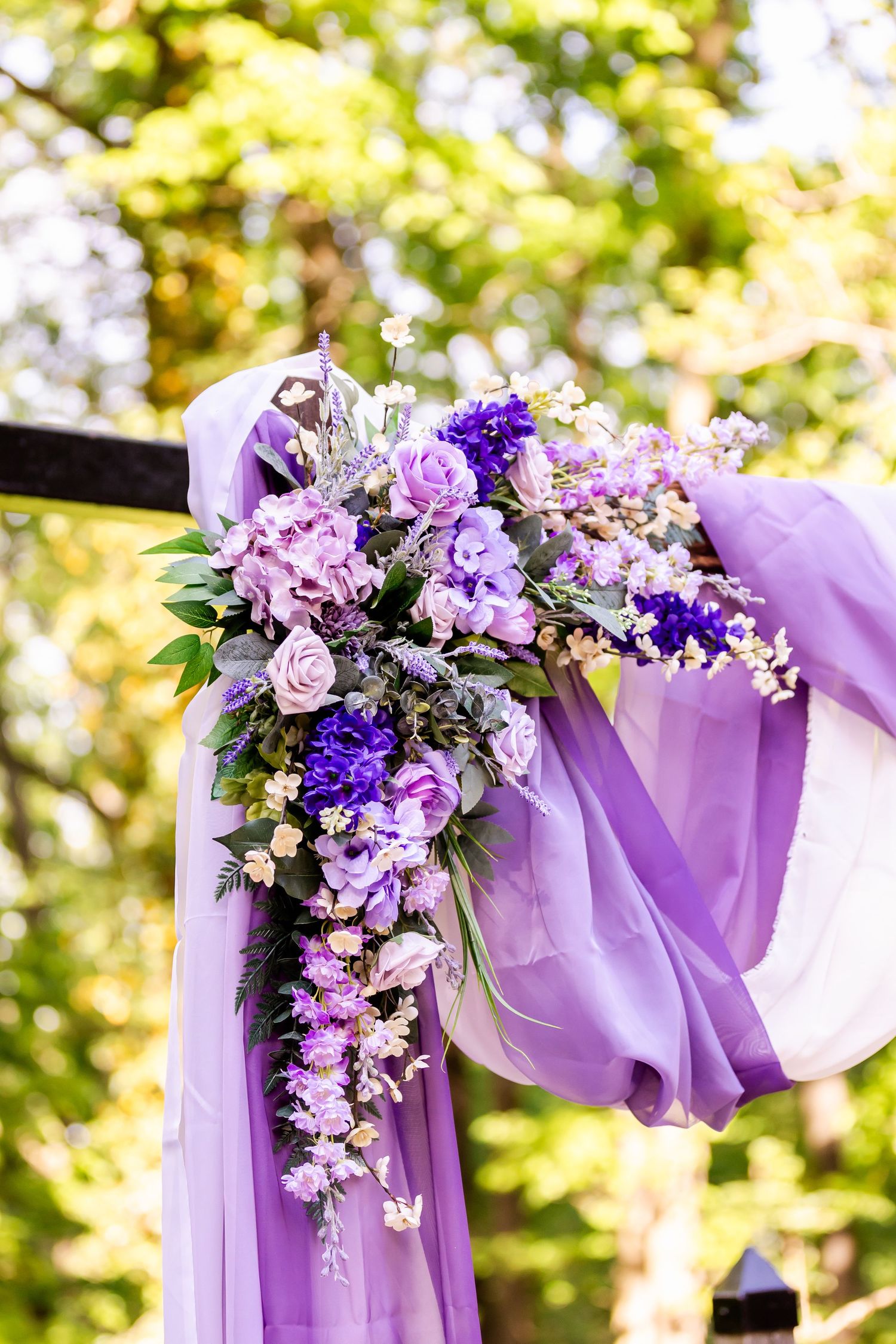 Purple and pink floral wedding arch decoration with flowing lavender fabric against a sunlit outdoor backdrop.